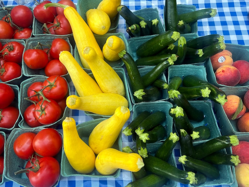 A photograph of a table with a blue gingham cloth full of blue produce boxes lined up with two rows of bright red tomatoes, yellow squash, zucchini and peaches.
