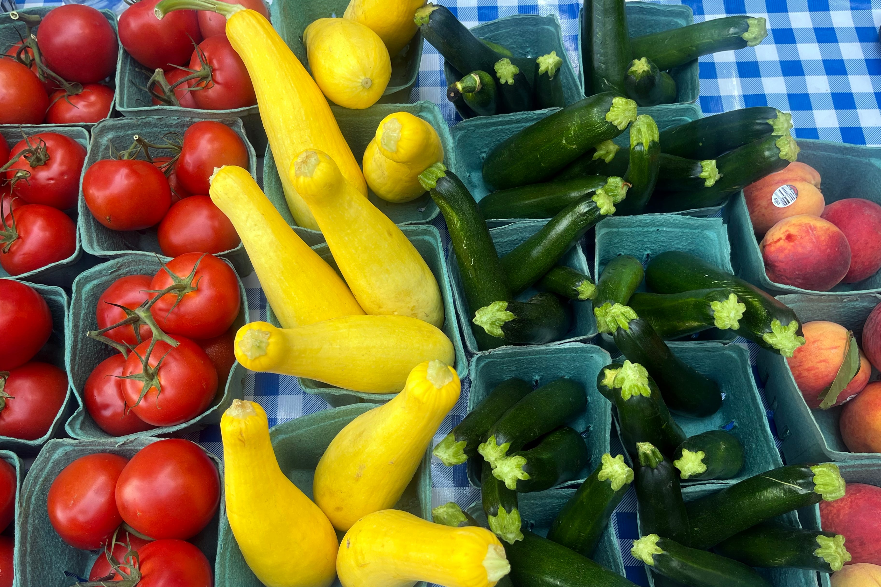 A photograph of a table with a blue gingham cloth full of blue produce boxes lined up with two rows of bright red tomatoes, yellow squash, zucchini and peaches.