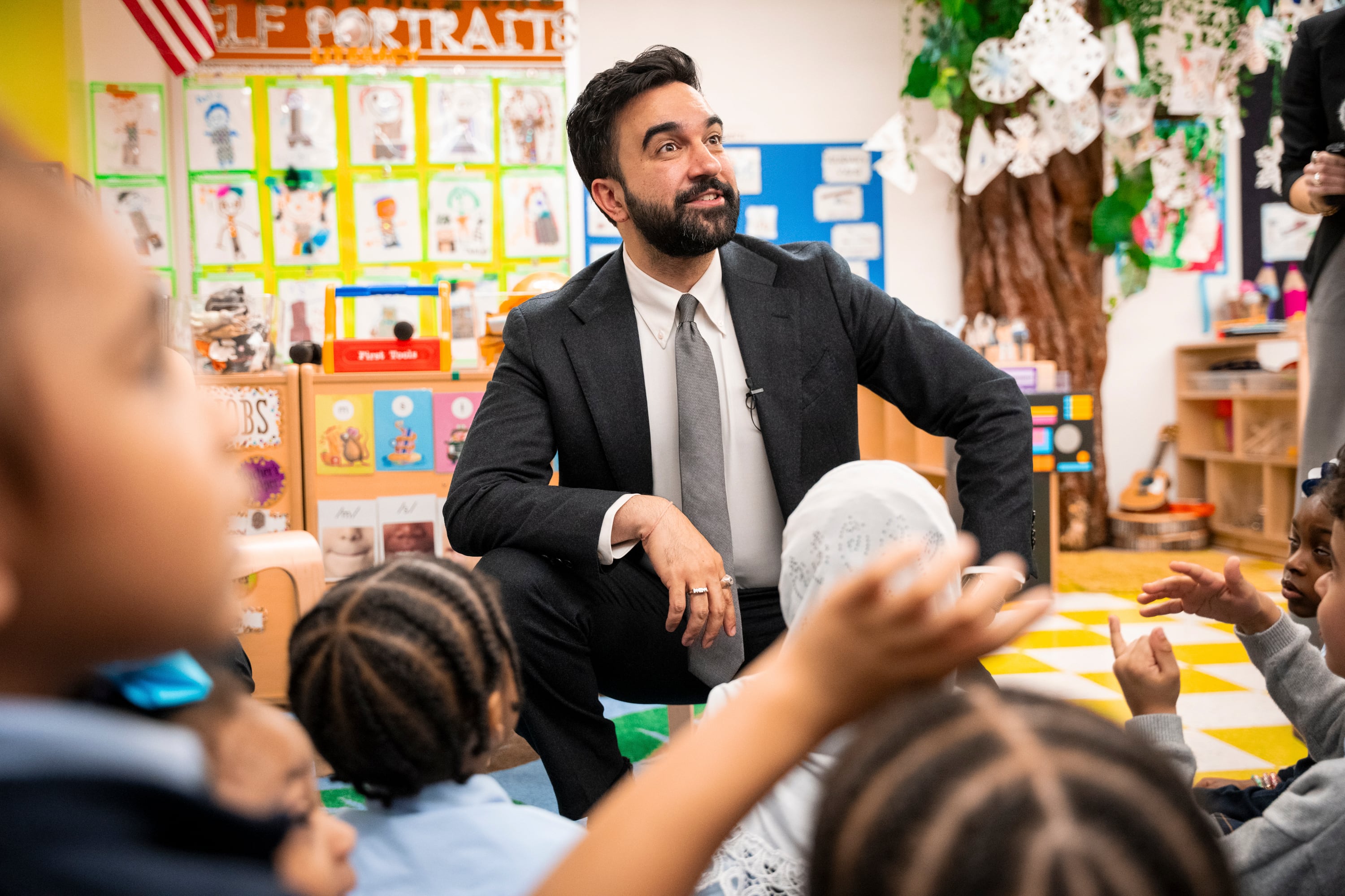 A light-skinned male is sitting in front of children and listening.
