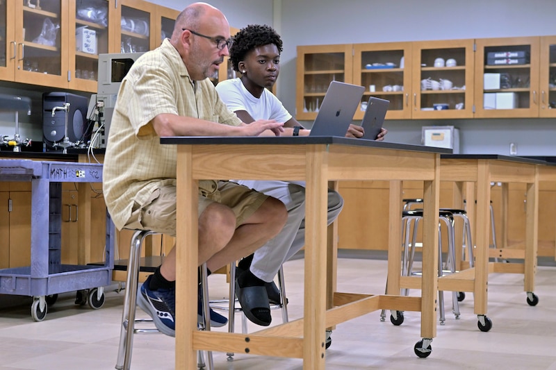 A teacher and a student sit next to each other at a table in a classroom.