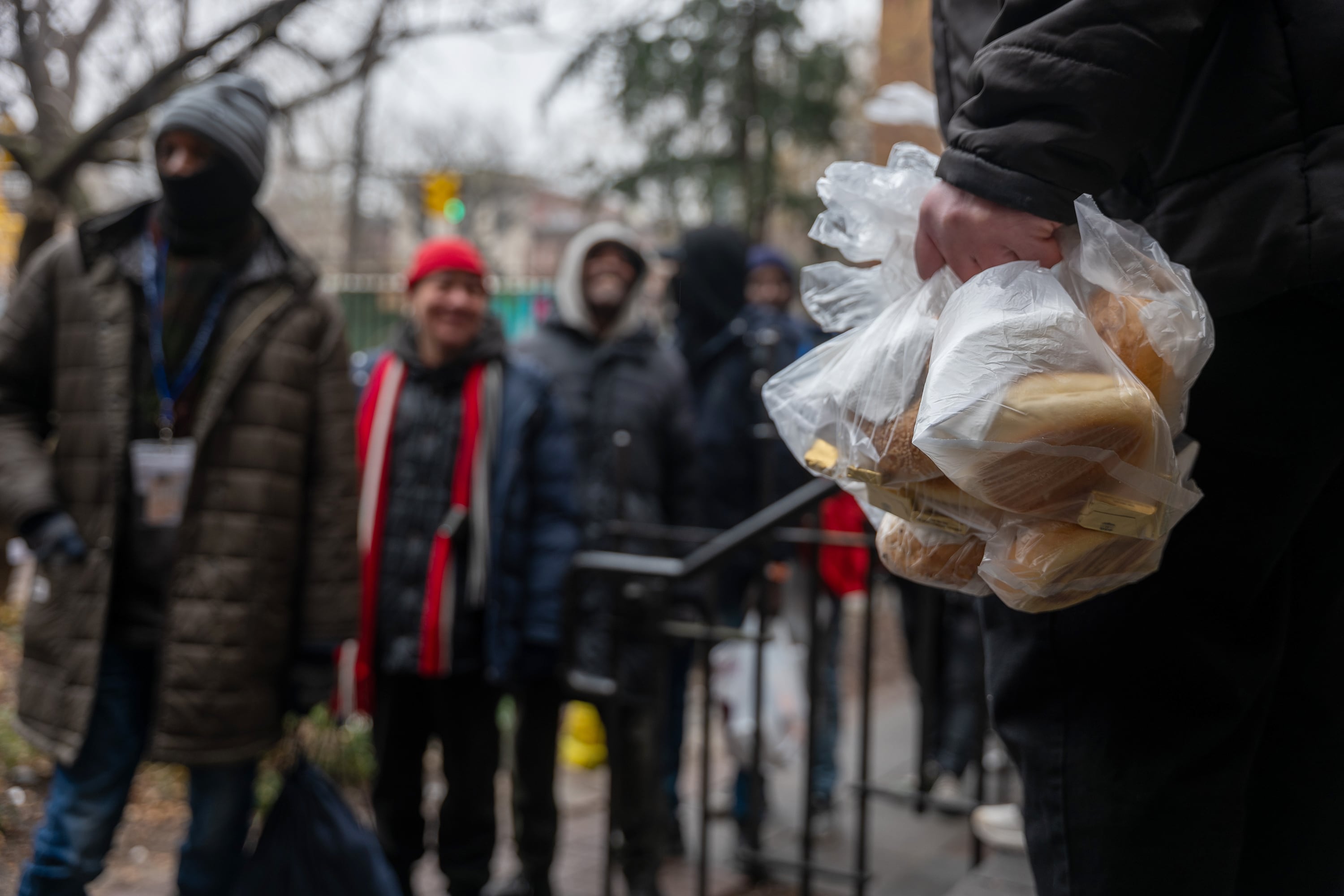 In focus is a hand holding bags full of bread with an out-of-focus line of people wearing winter clothing.