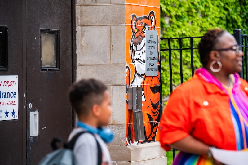 A Black woman in a colorful dress speaks to a young student outside of a school building.