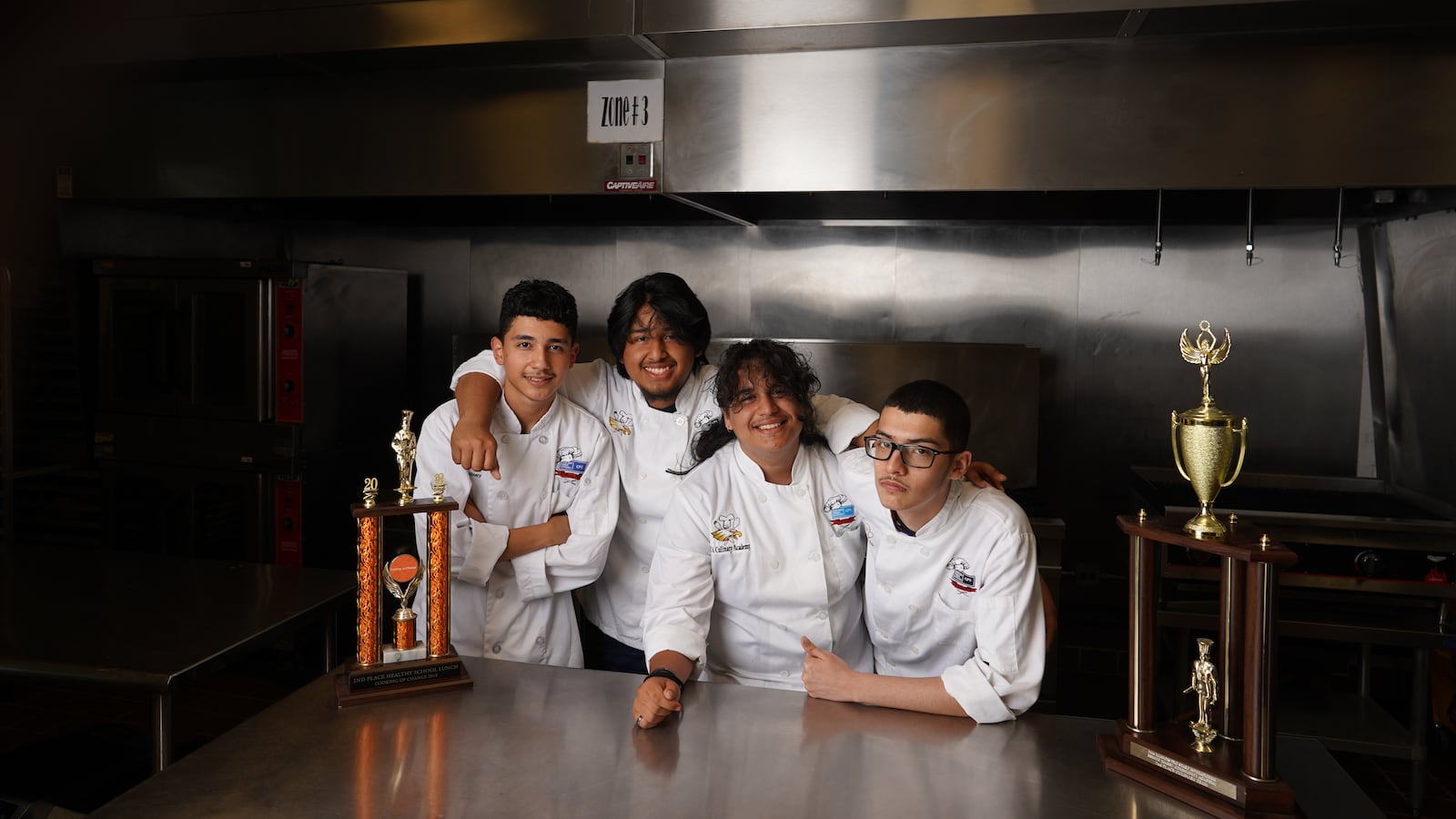 Four culinary students, all wearing white coats, pose on a prep station together with two large trophies.