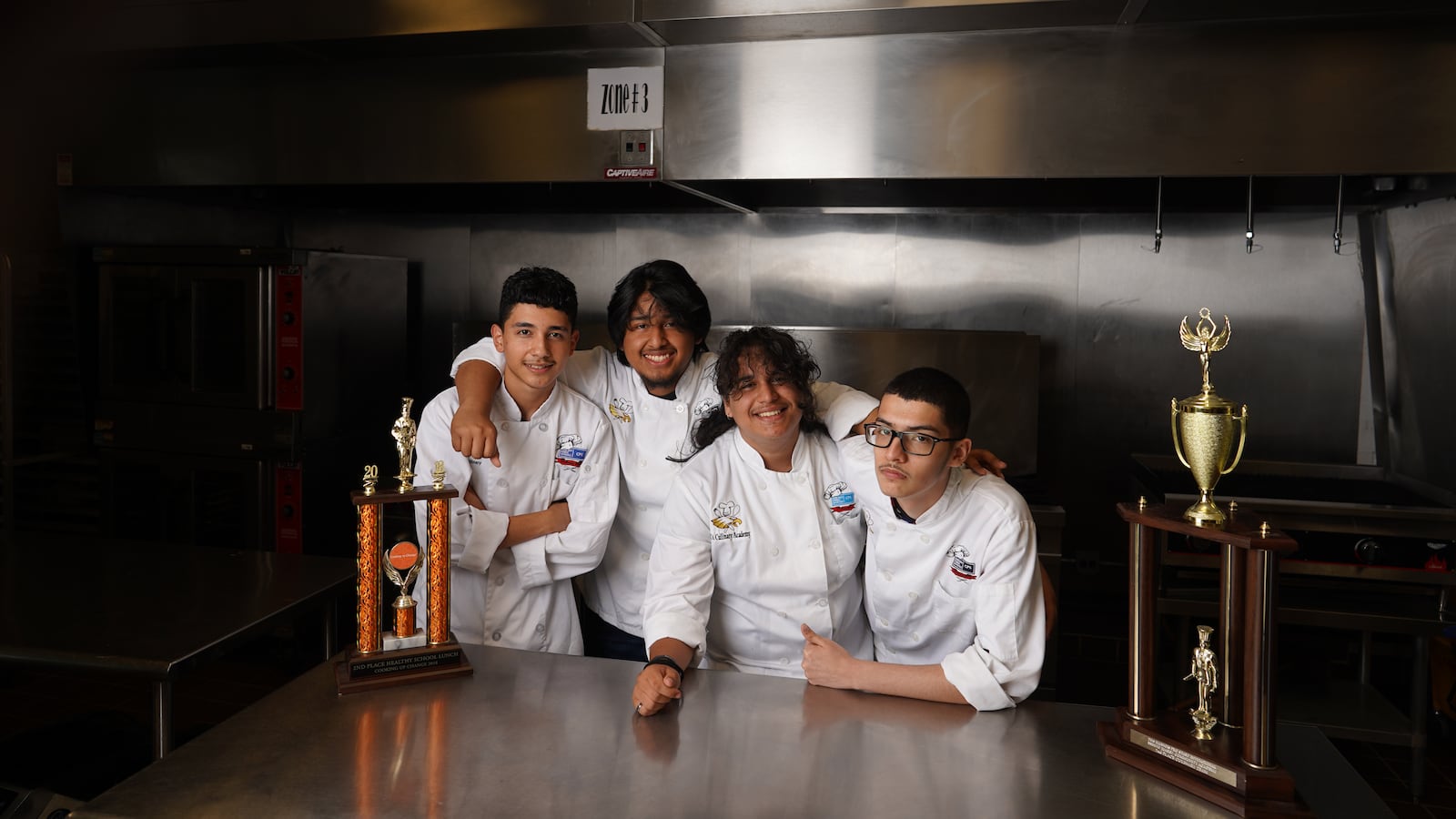 Four culinary students, all wearing white coats, pose on a prep station together with two large trophies.