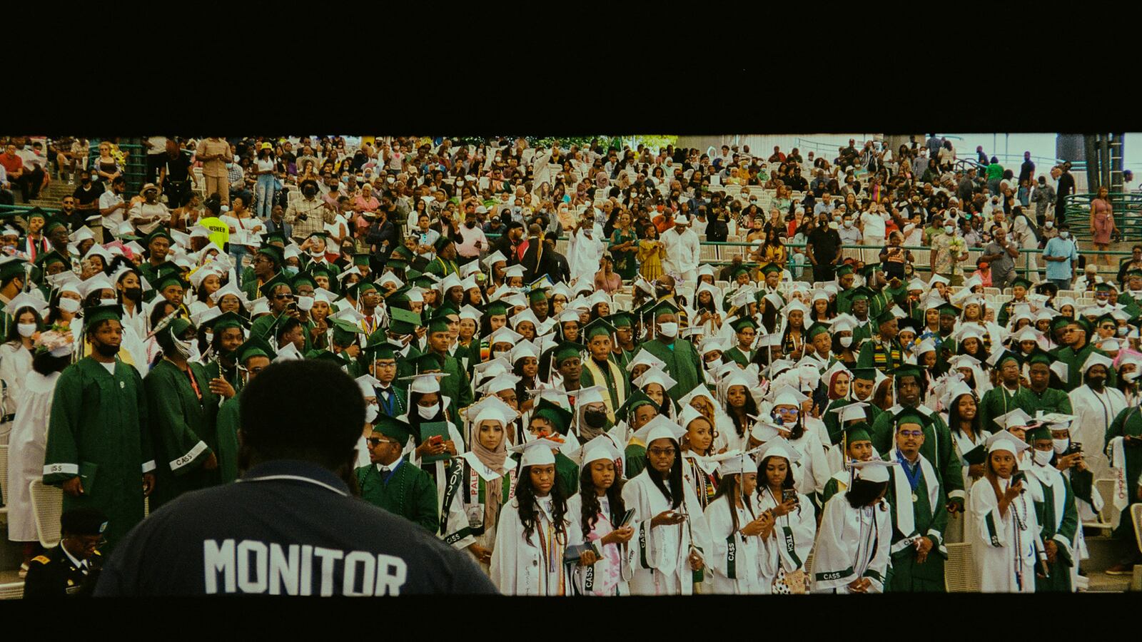 A large group of graduating high school students, wearing green and white caps and gowns, stand in neat rows as their parents look on behind them.