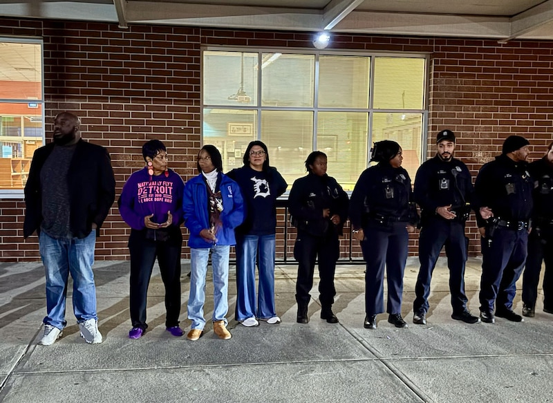 A photograph of a group of adults and some police officers standing in a line outside of a school building waiting to cheer on student.