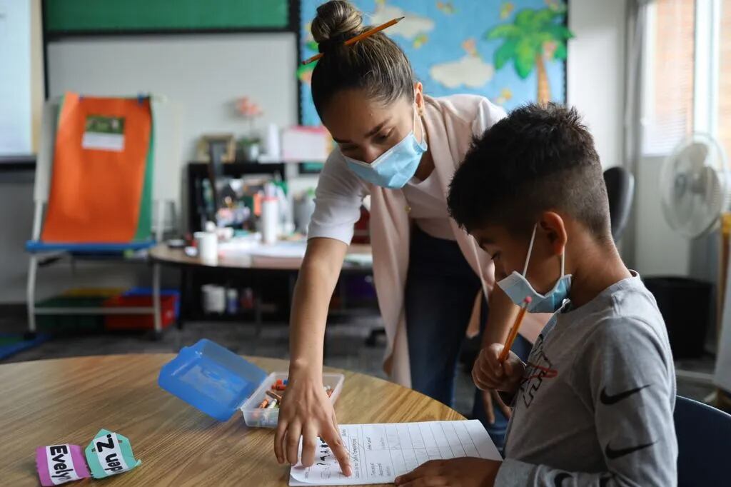 An adult helps a young student with a worksheet in a classroom. Both are wearing masks.