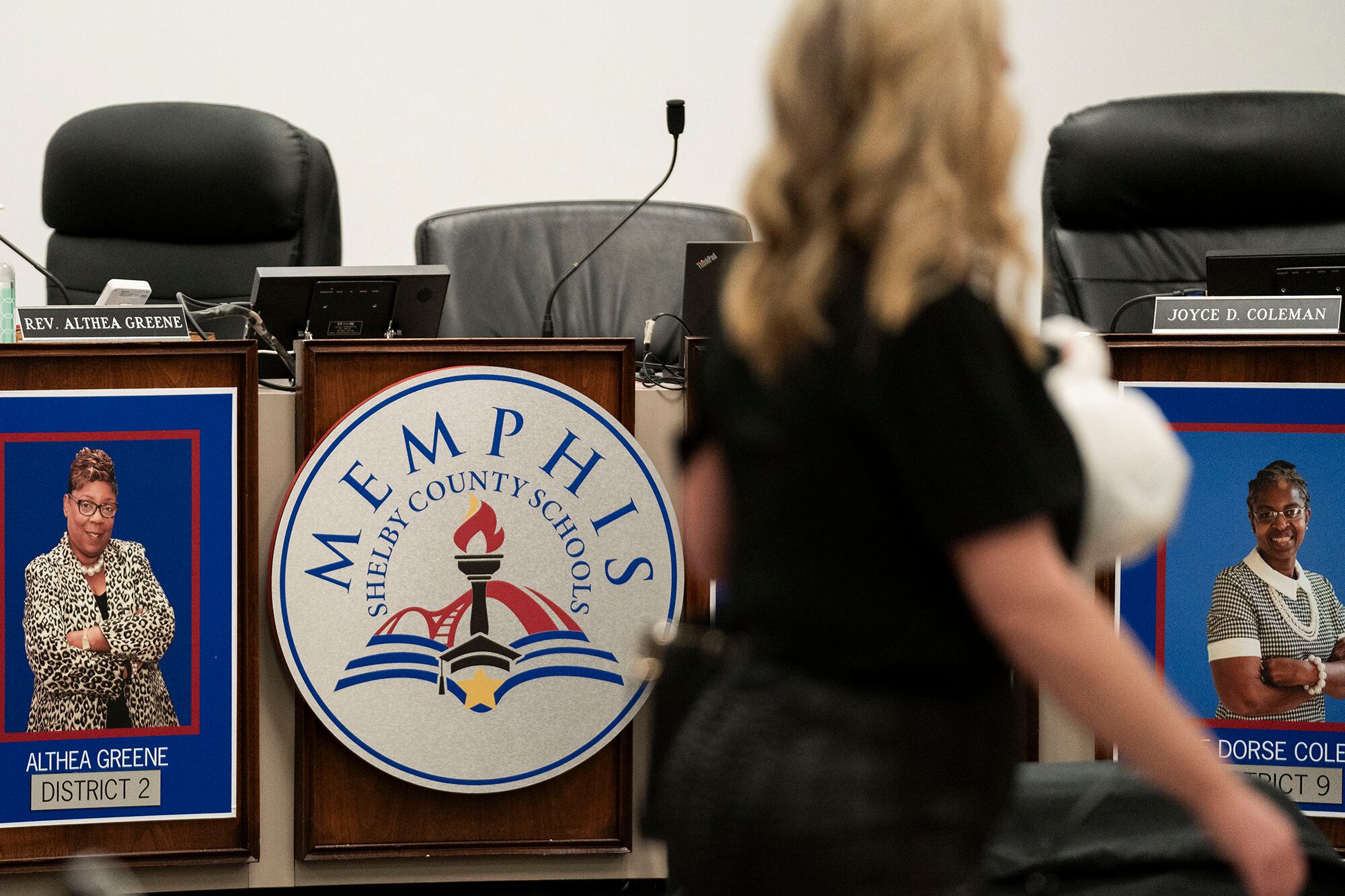 A woman wearing a black shirt walks in front of a desk with chairs in the background. There is a large logo on the side of the table that reads, "Memphis Shelby County Schools."