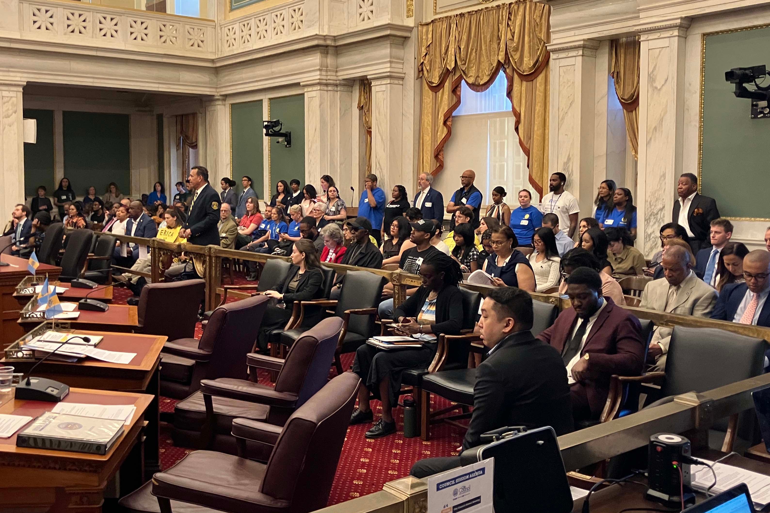 A photograph of a large group of people sitting in a large city council room.