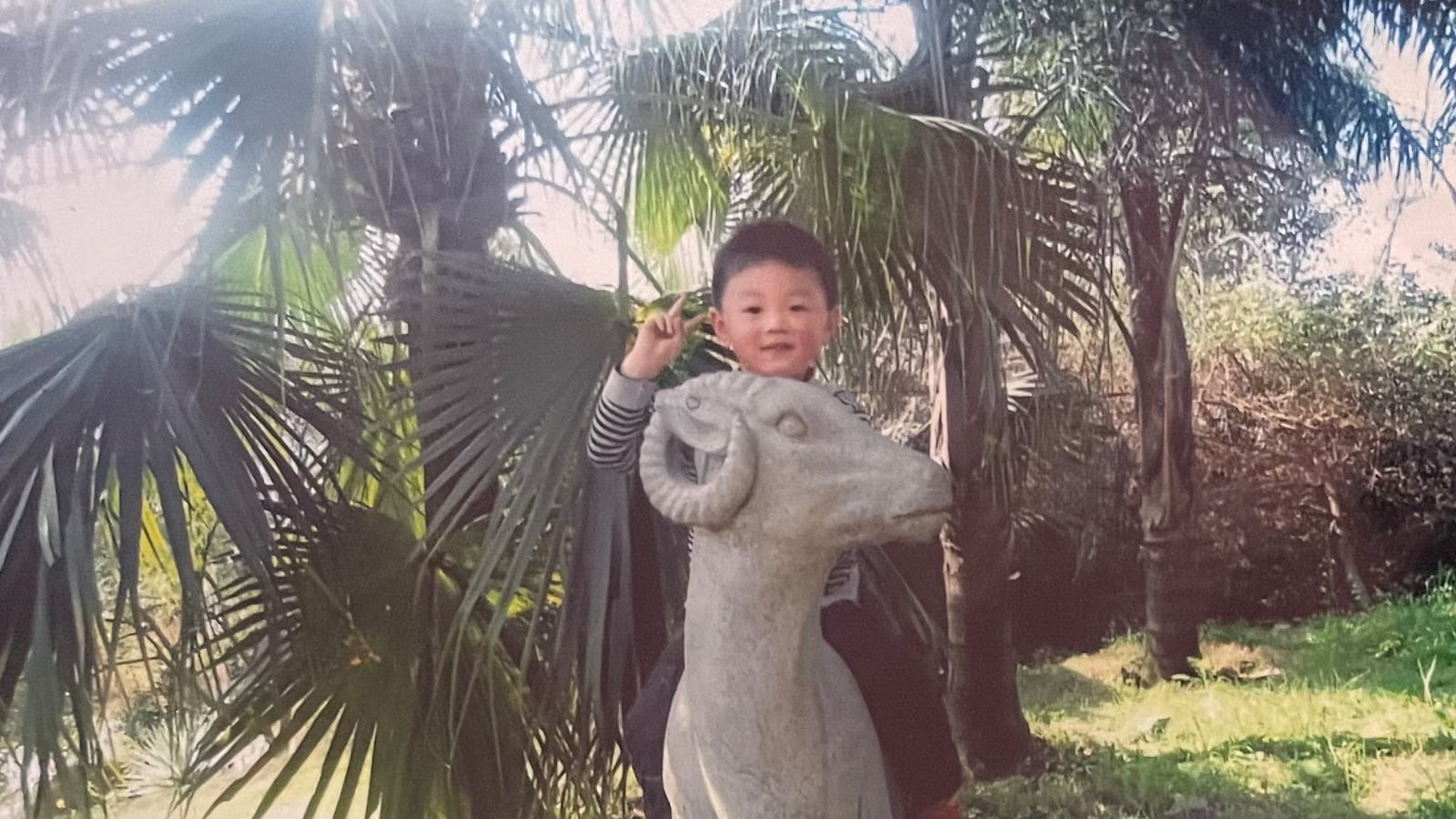 Photo of a young boy sitting on a statue of a ram. There are palm trees behind him.