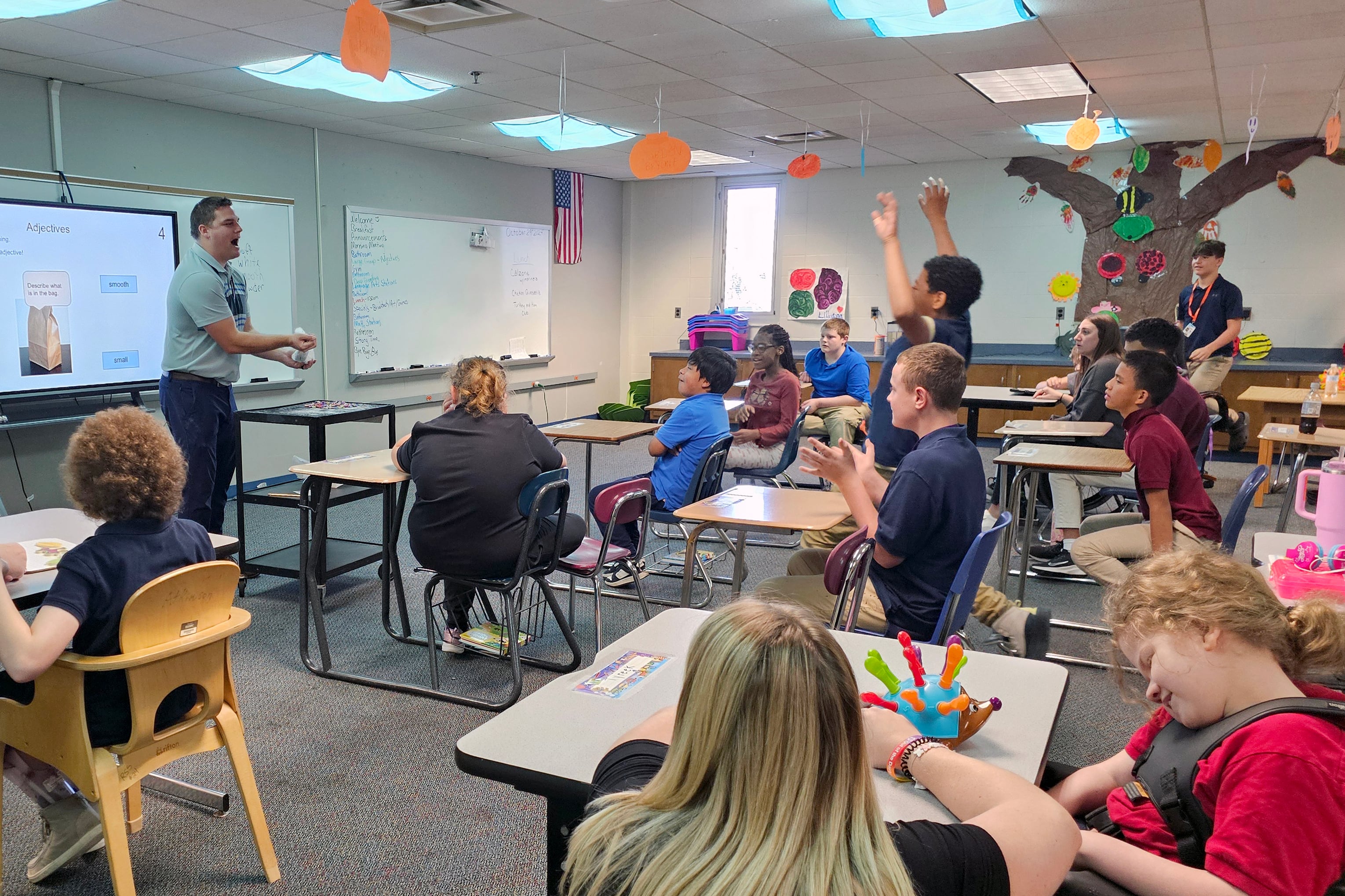 An adult man wearing a blue shirt teaches from the front of a classroom to a room full of students sitting at desks.
