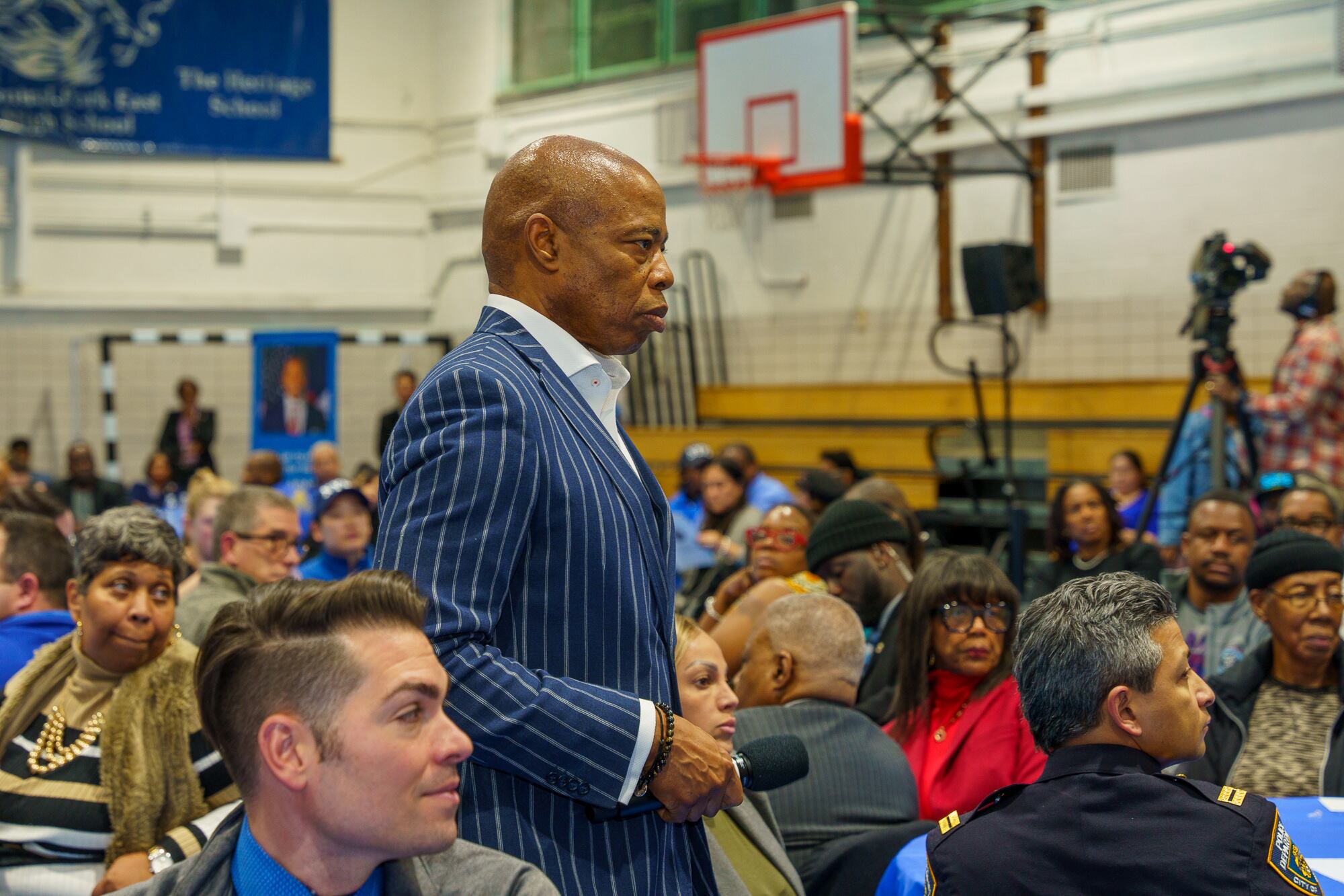 A man wearing a blue striped suit stands in a school gym full of people with a basketball hoop in the background.