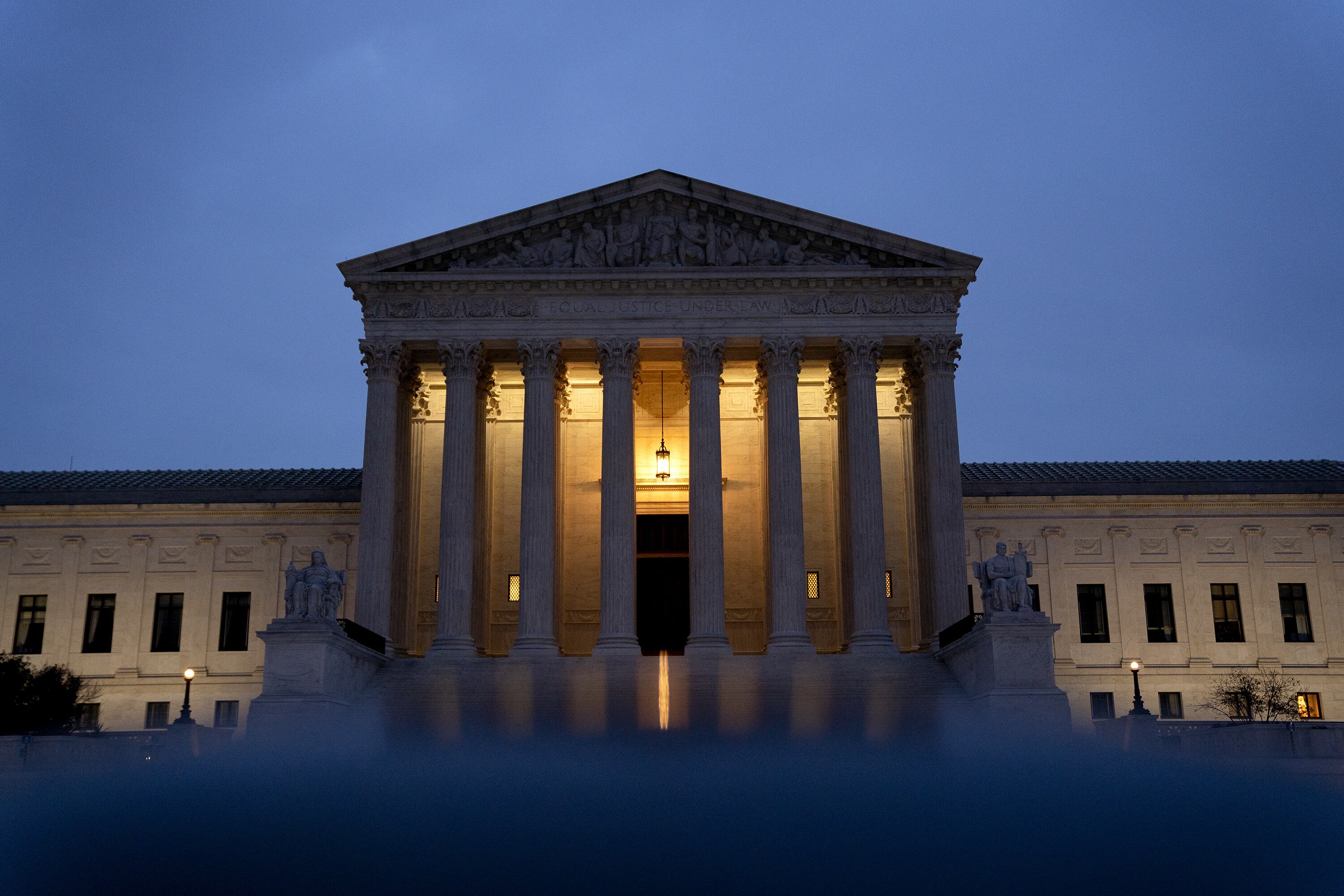 The U.S. Supreme Court building with large stone pillars at the front entrance with a dark blue sky in the background. The lights of the building are illuminating while the sun either sets or rises.