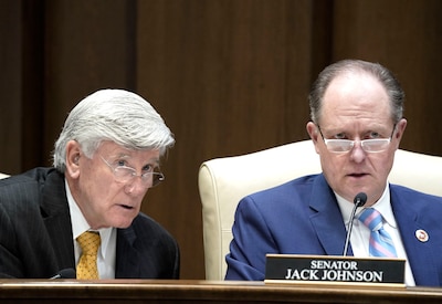 Two men look on with their mouths open while they sit at a wooden desk with wooden paneling in the background.