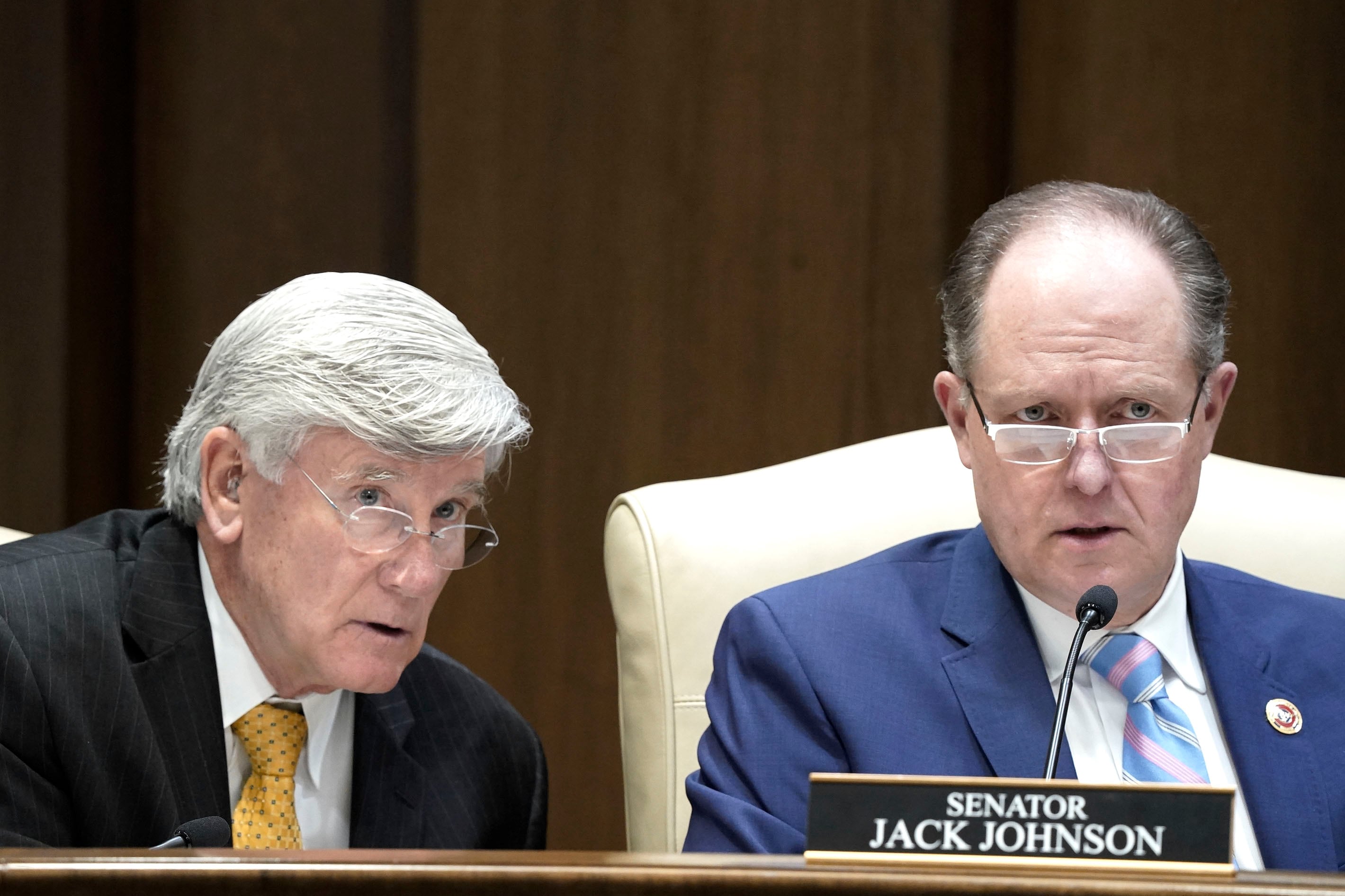 Two men look on with their mouths open while they sit at a wooden desk with wooden paneling in the background.