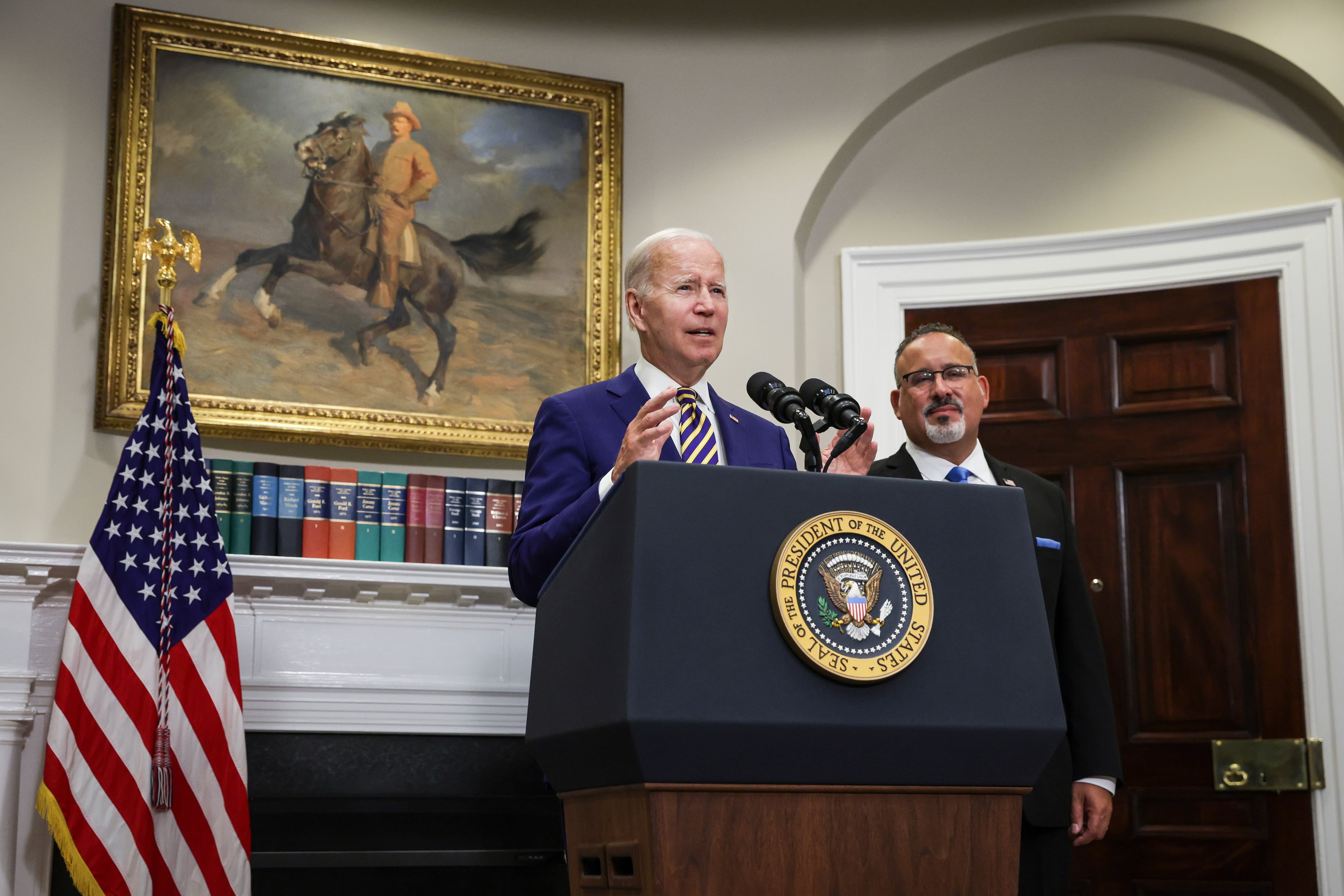 President Joe Biden speaks from a podium while Education Secretary Miguel Cardona stands to the right.
