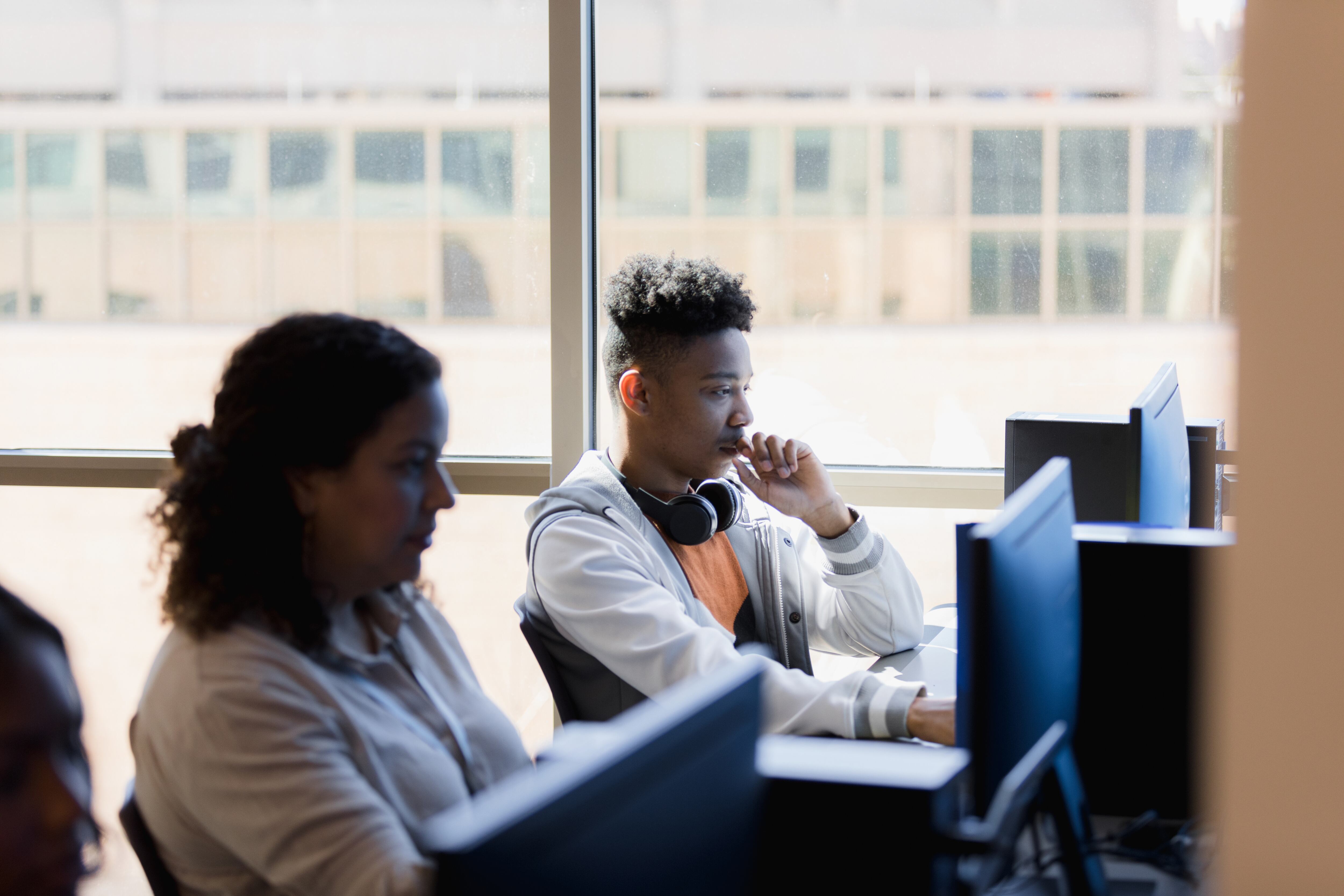 Two students sit in a classroom looking at laptop screens on a desk. There are charts on the wall in the background.