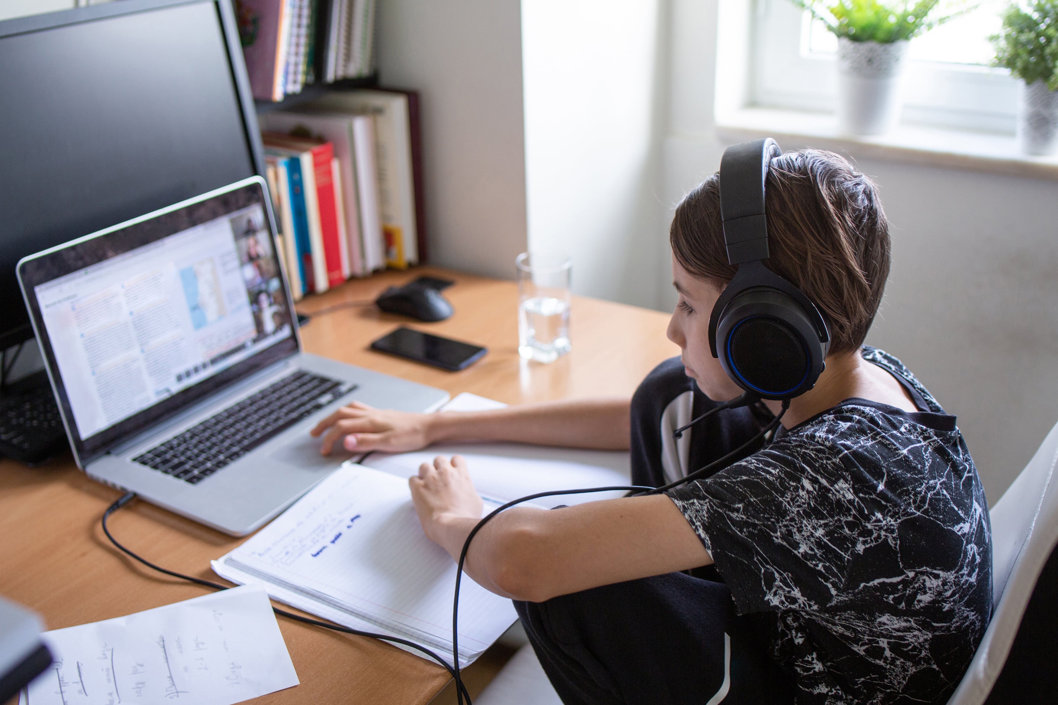 A young student works at a desk in front of a laptop while waring large head phones.