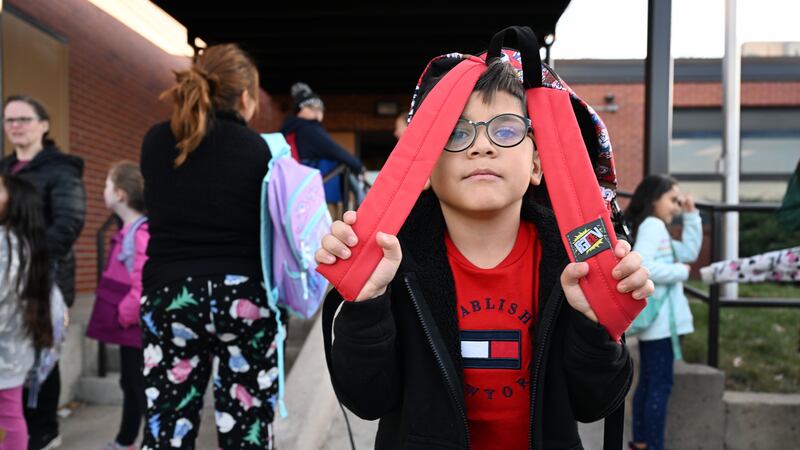 A boy with glasses peeks out from behind his backpack straps while he waits outside his school.