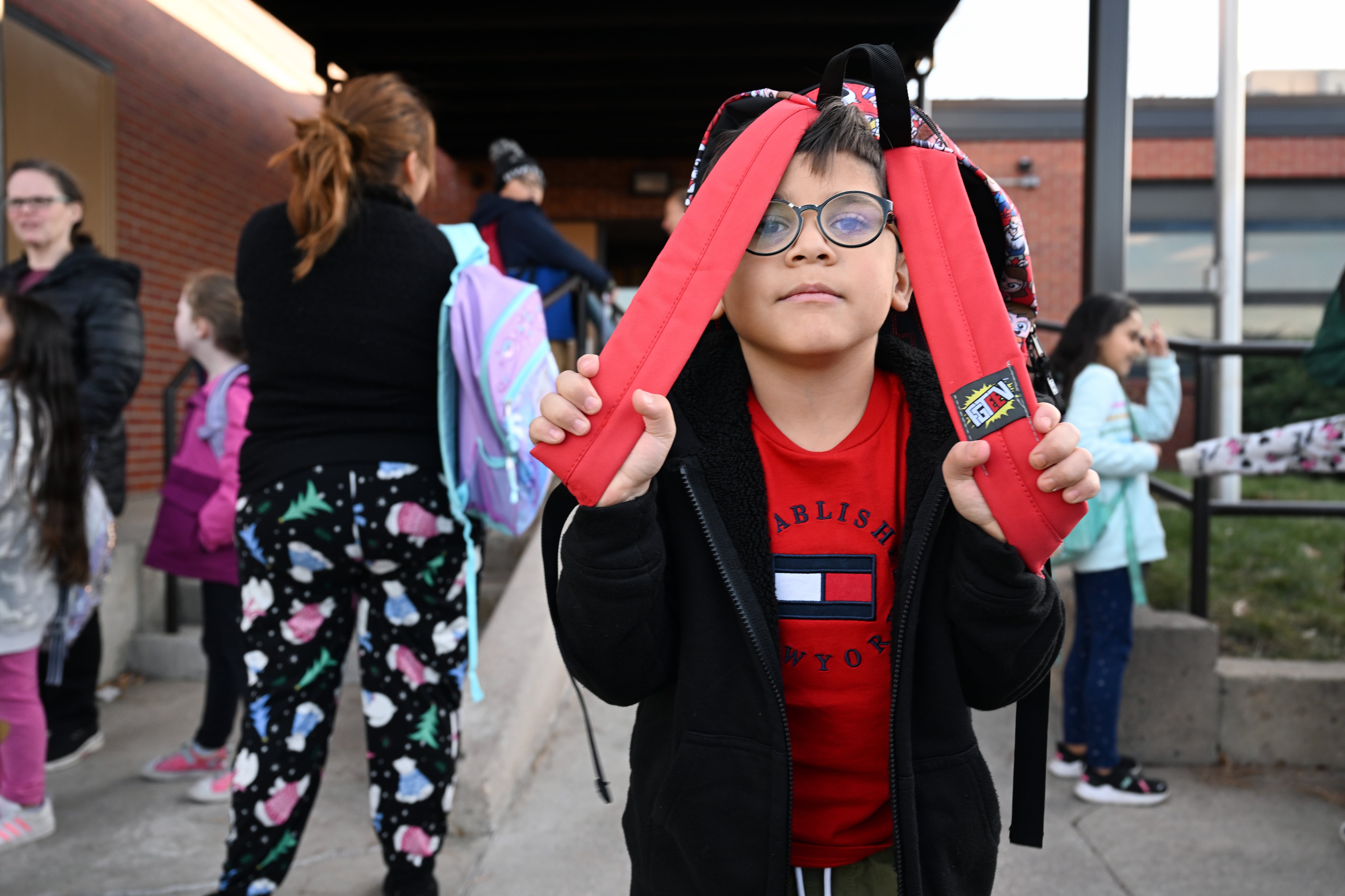 A boy with glasses peeks out from behind his backpack straps while he waits outside his school.