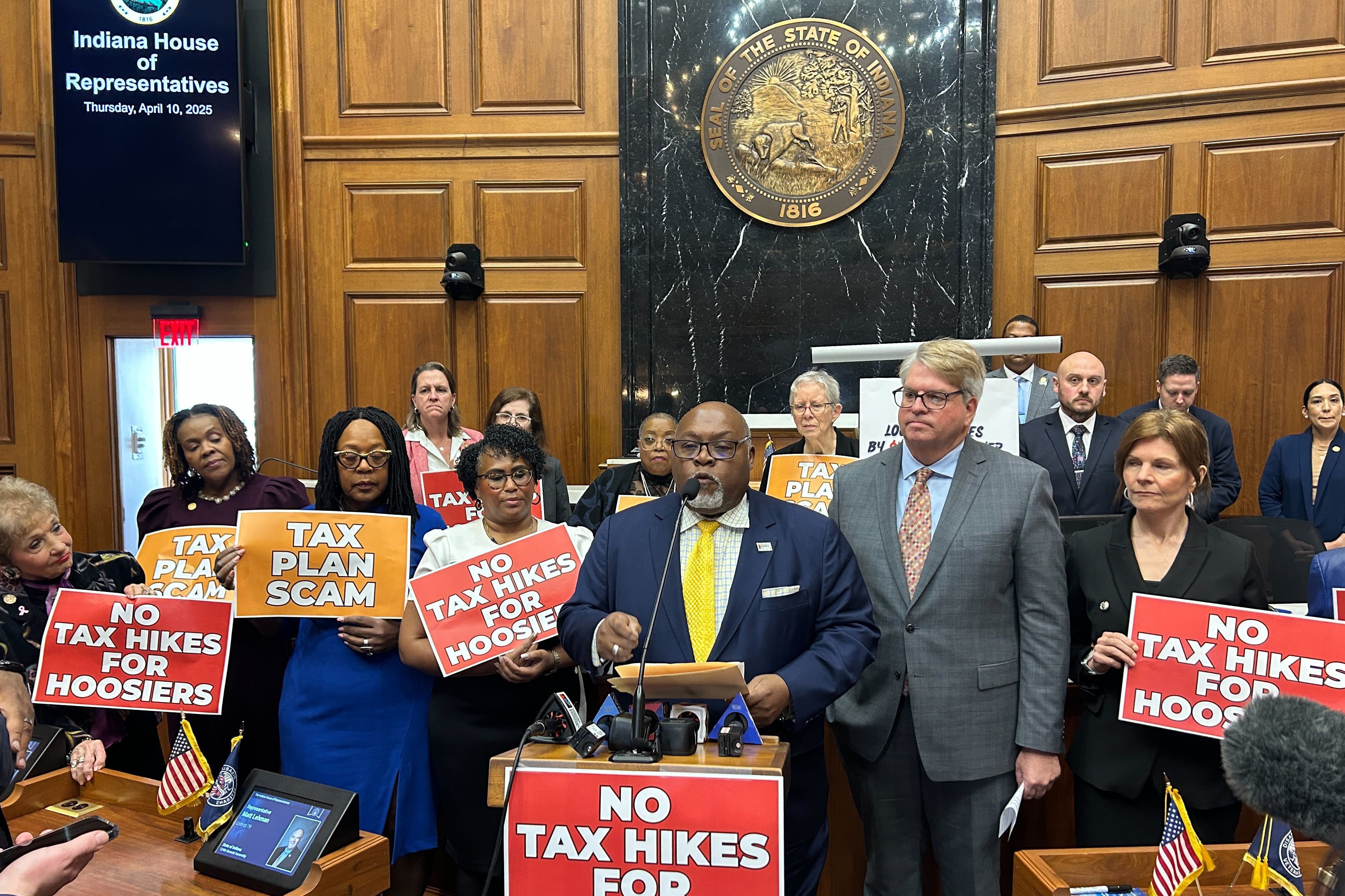 A large group of people in suits and business clothing stand behind a podium in a large Capitol room. Some of the people are holding signs that read "No Tax Hikes For Hoosiers."