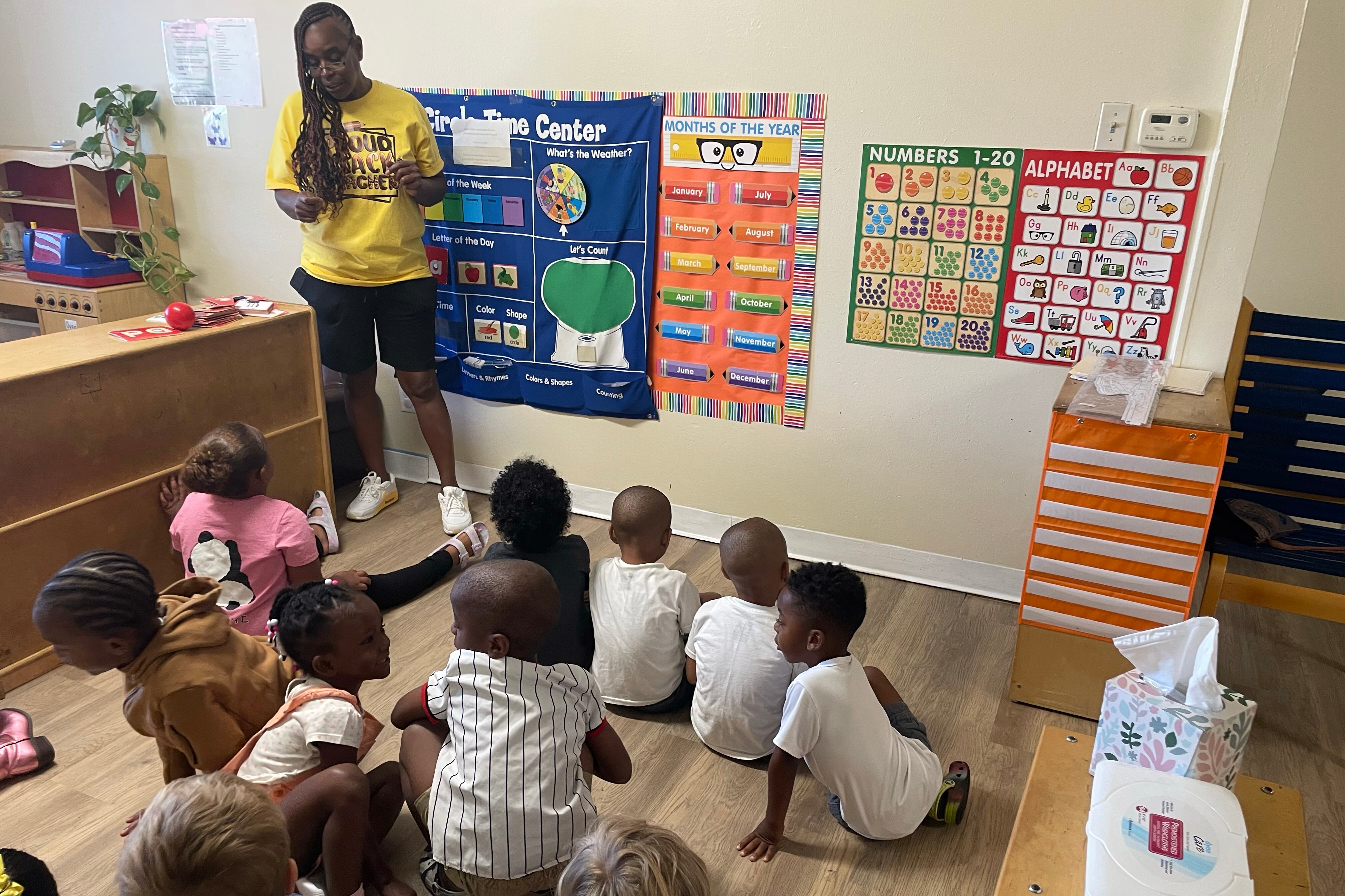 A teacher with long dark hair and wearing a yellow t-shirt stands in front of a classroom full of young students sitting on the floor. There are colorful posters on the wall in the background.