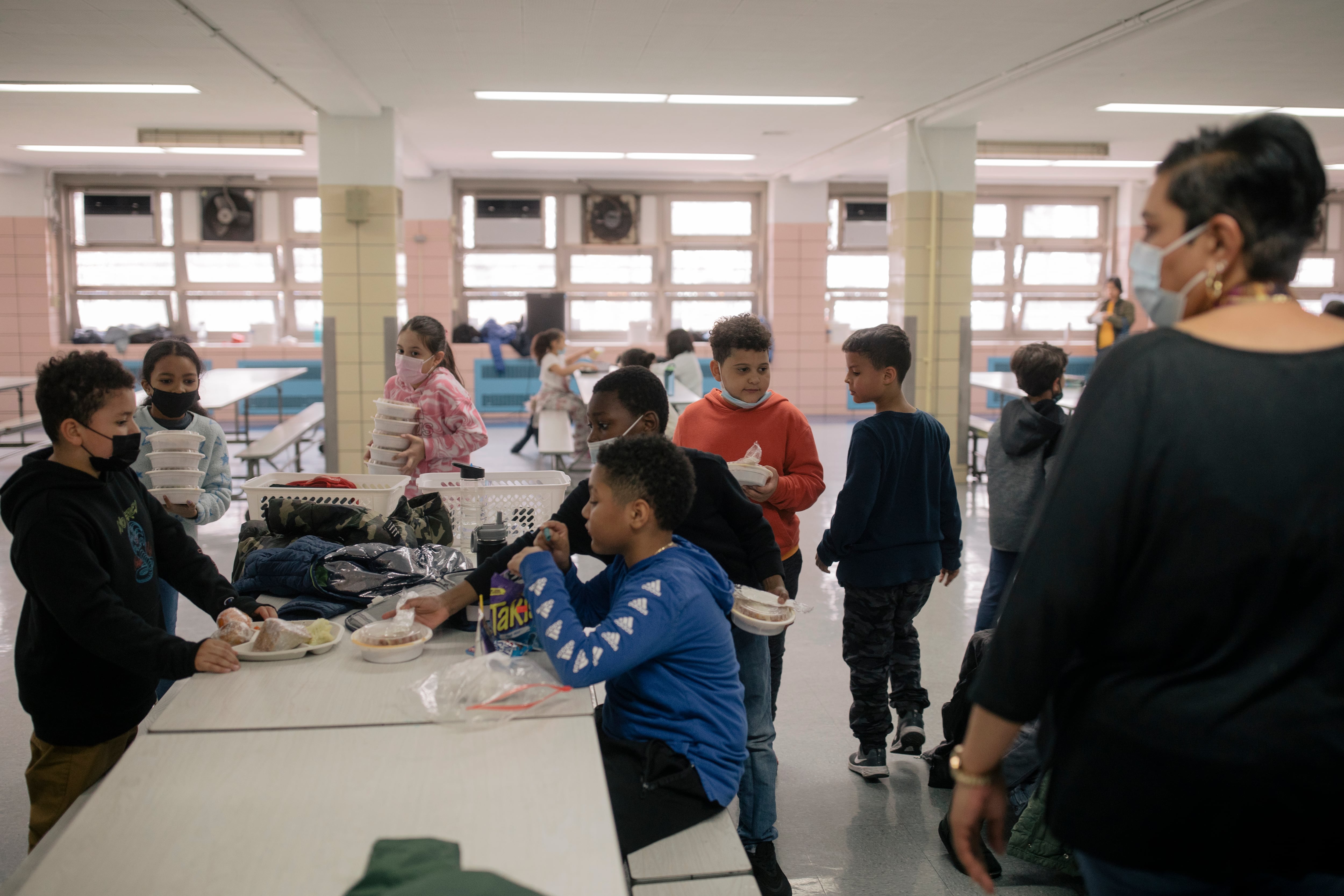 Ten students are scattered across a cafeteria, with five eating lunch at a table as an adult stands nearby.
