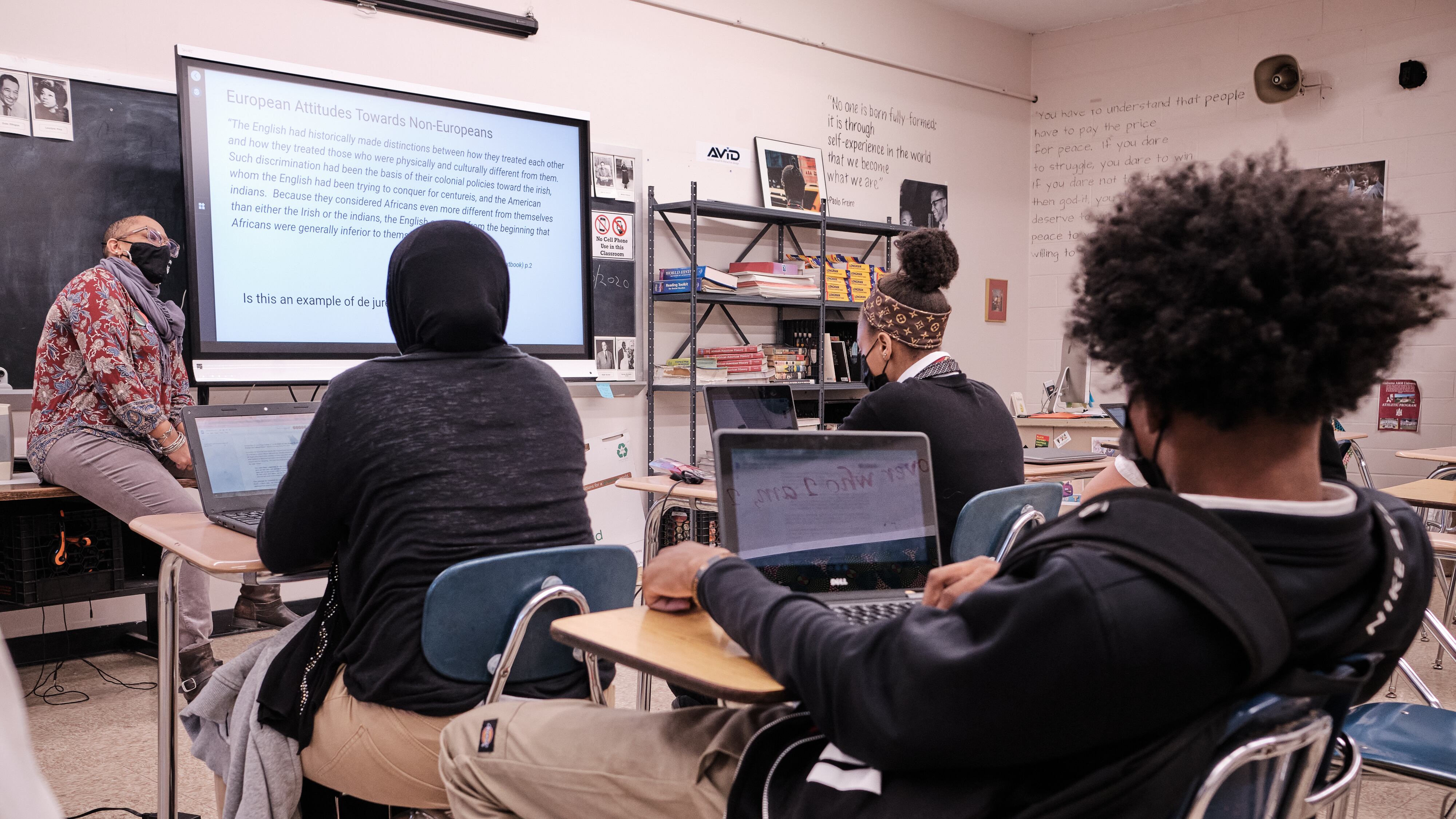 A woman wearing a black face mask and glasses sits on a table in front of several students in a classroom. To her left is a projector screen that reads “European Attitudes Towards Non-Europeans.”