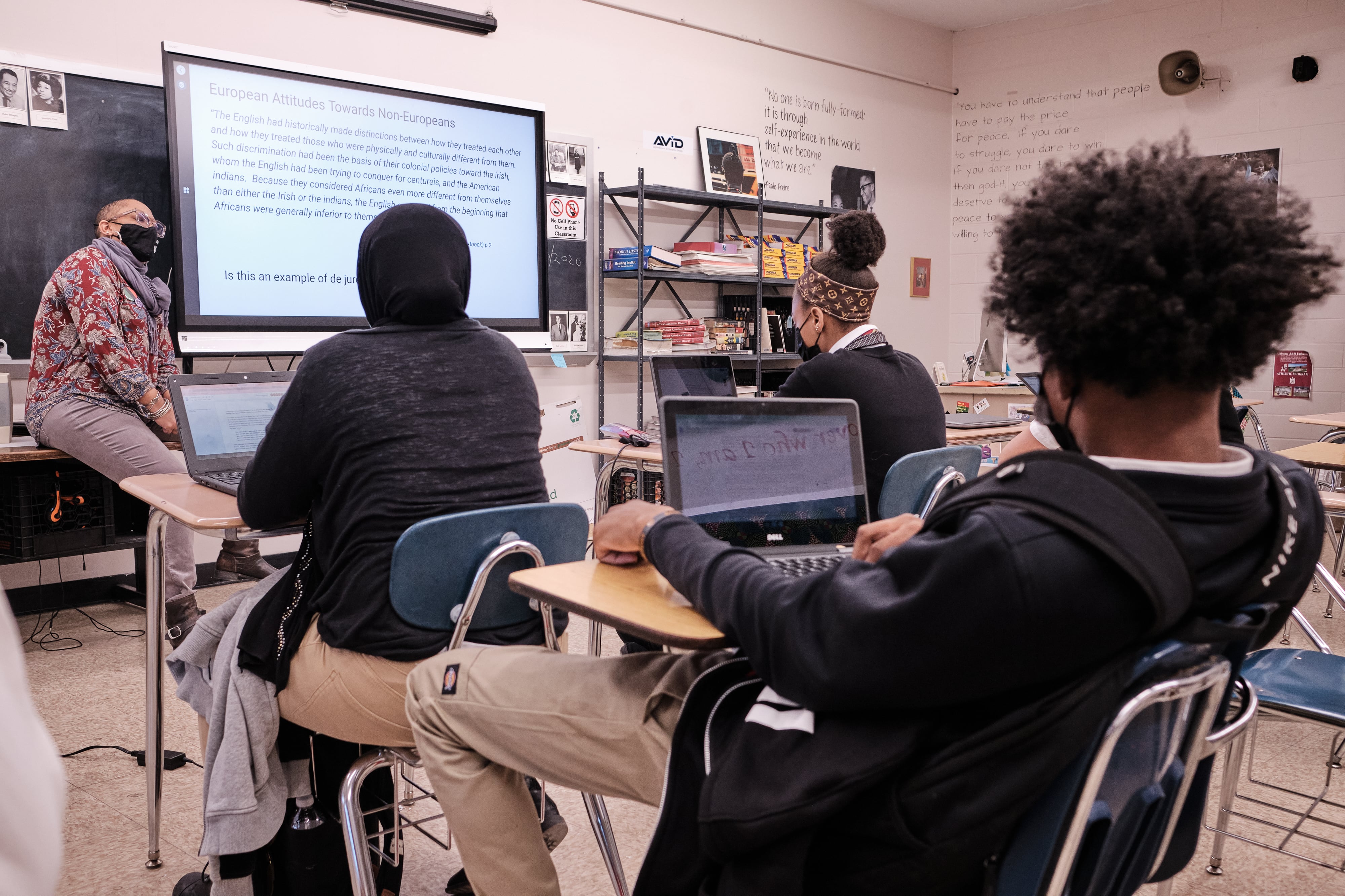 A woman wearing a black face mask and glasses sits on a table in front of several students in a classroom. To her left is a projector screen that reads “European Attitudes Towards Non-Europeans.”