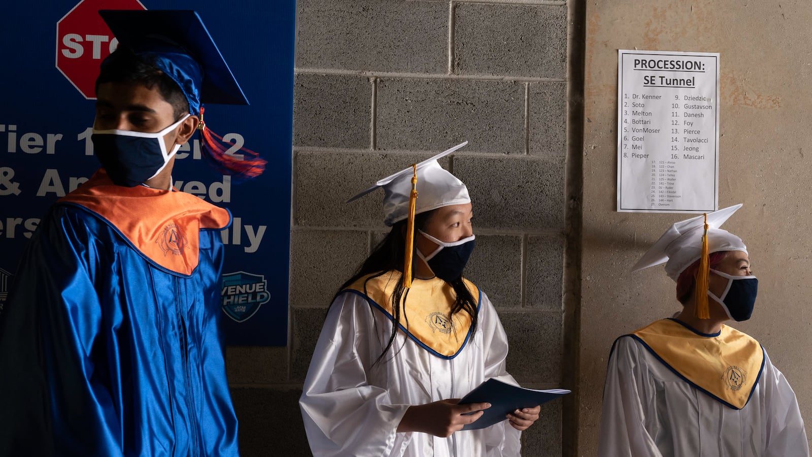 A young man in blue and orange graduation regalia stands next to two young women wearing white and gold graduation caps and gowns, each holding programs in their hands.