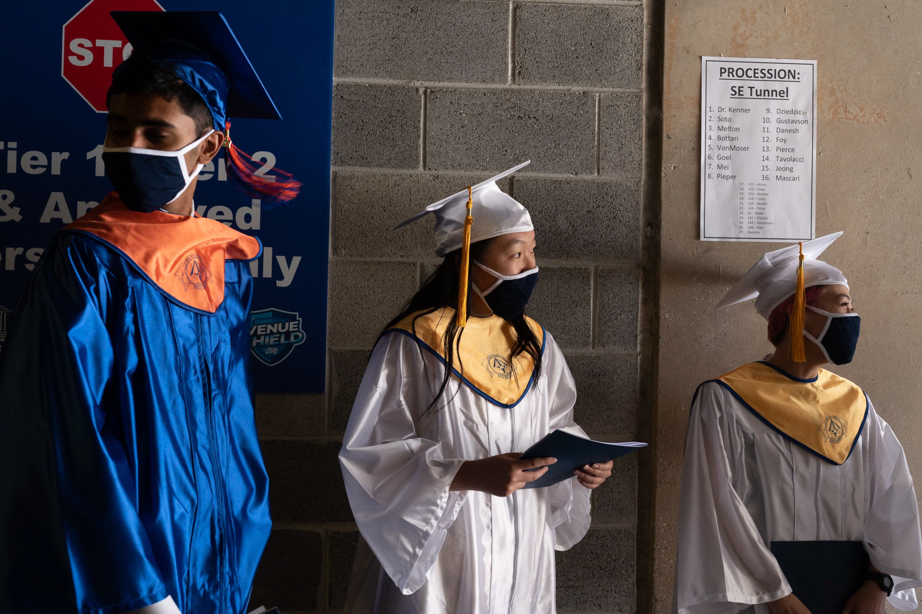 A young man in blue and orange graduation regalia stands next to two young women wearing white and gold graduation caps and gowns, each holding programs in their hands.
