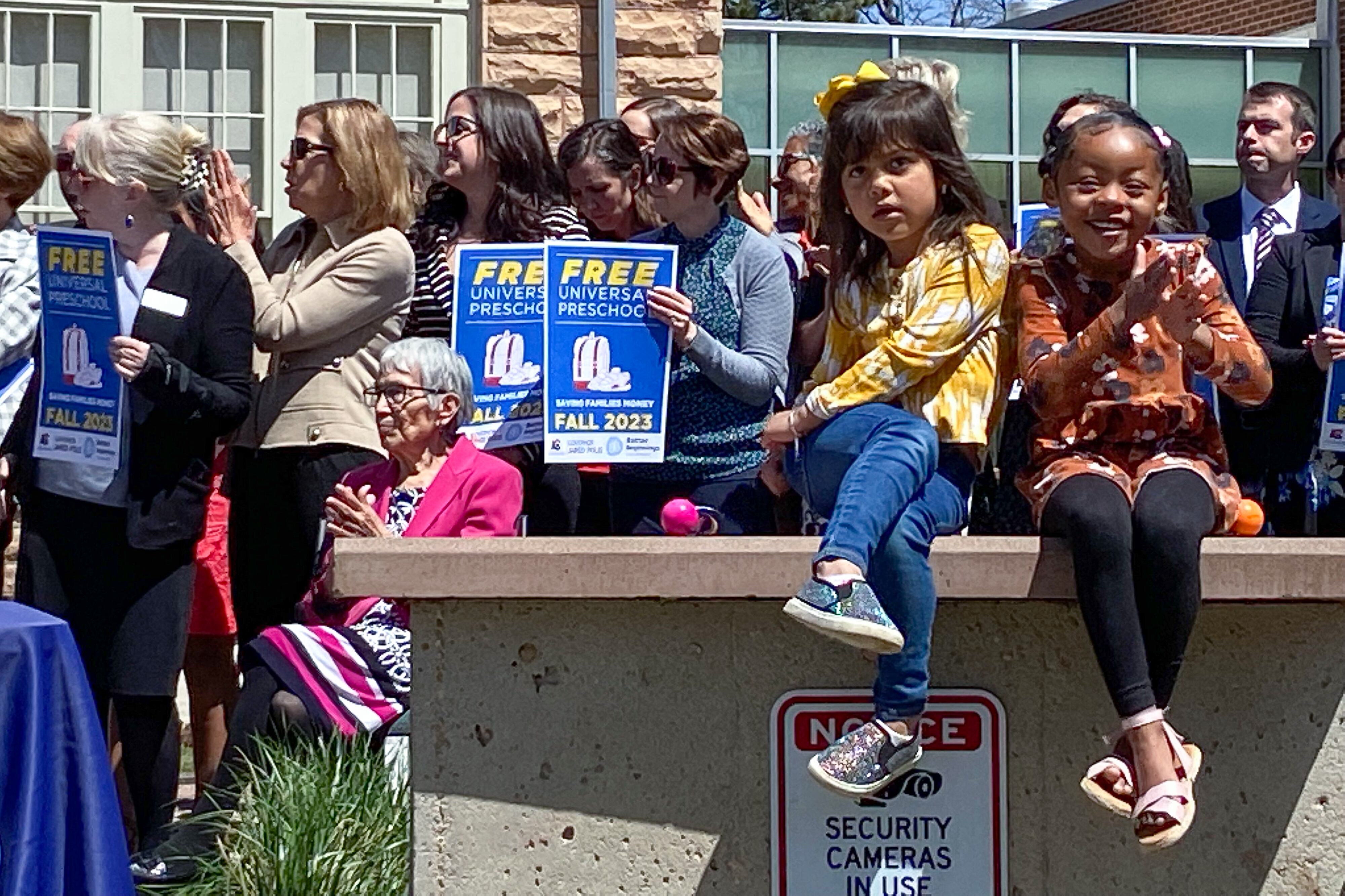 Two little girls sit on a brick wall. One looks distracted and squirmy, while the other claps her hands. A large crowd of adults stands behind them. The adults hold signs that say “Free Universal Preschool.”