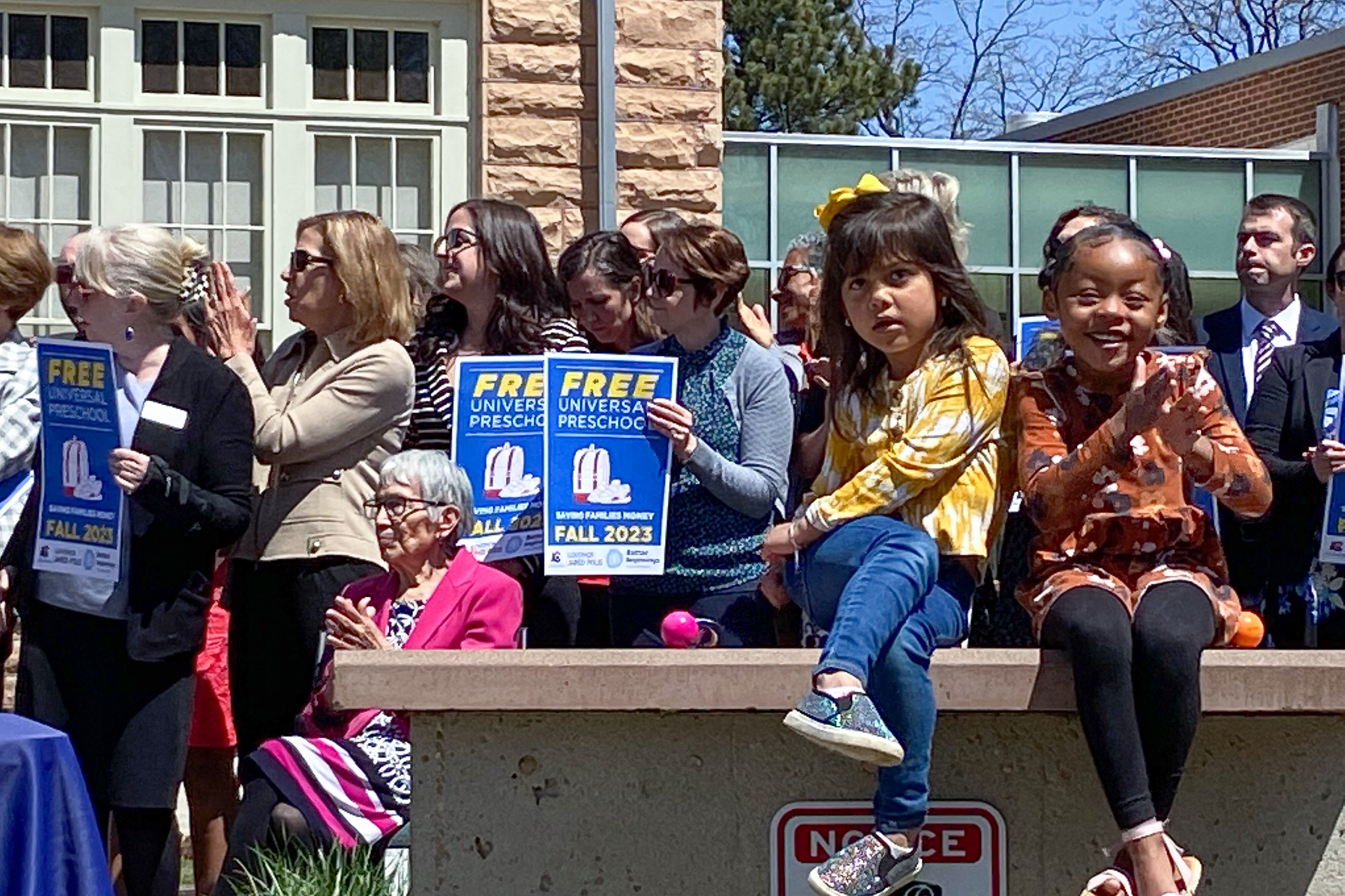 Two little girls sit on a brick wall. One looks distracted and squirmy, while the other claps her hands. A large crowd of adults stands behind them. The adults hold signs that say “Free Universal Preschool.”