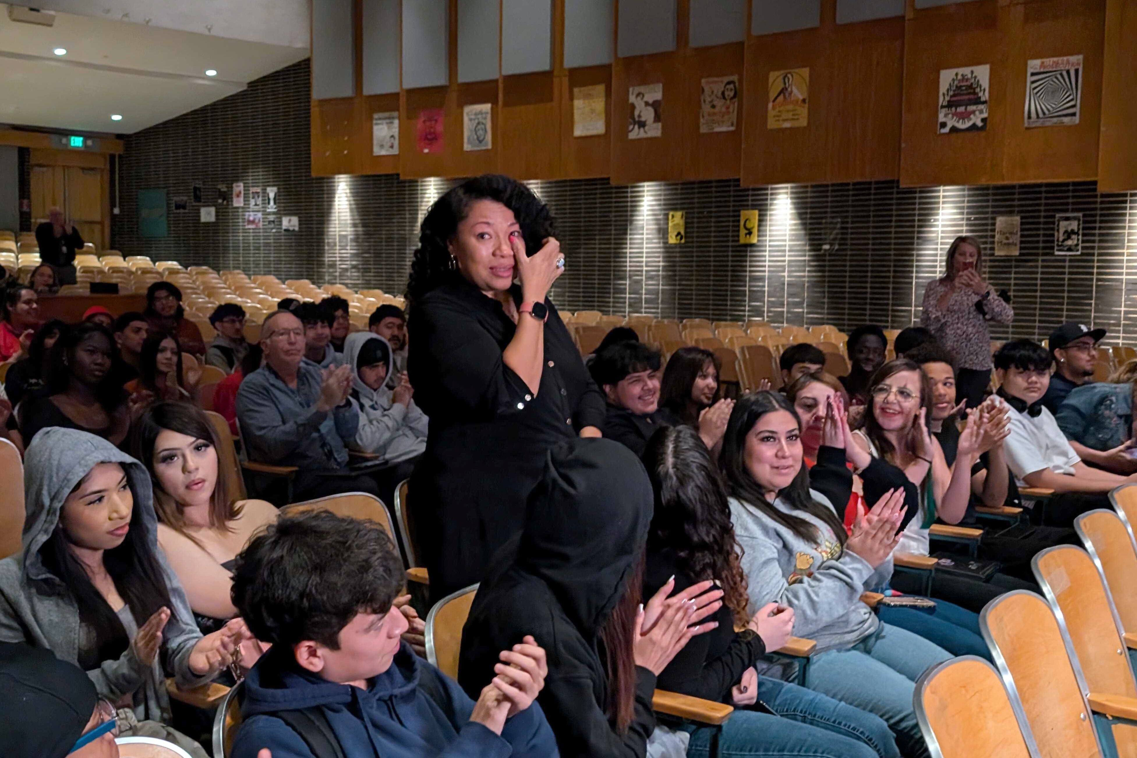 A woman wearing a dark dress wipes a tear away while standing in a row of chairs full of people in an auditorium.