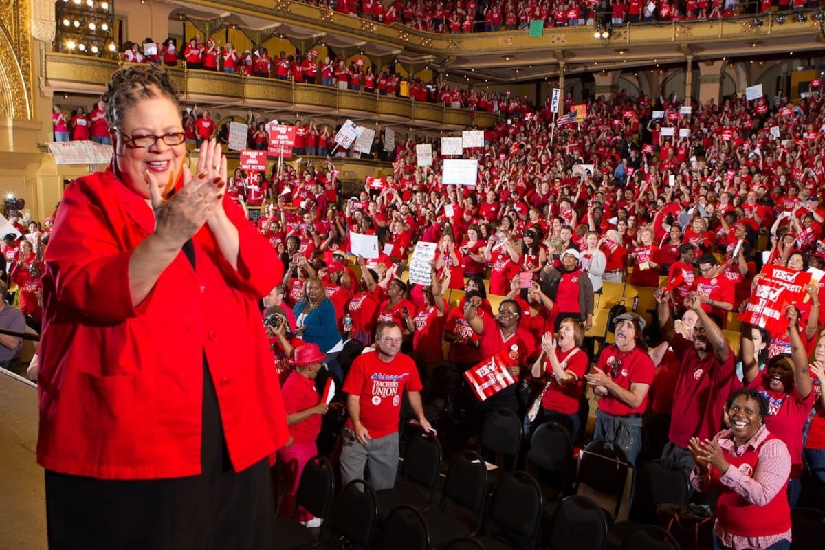 A Black woman clapping her hands and wearing a red shirt stands on stage near a large crowd of people wearing red shirts.