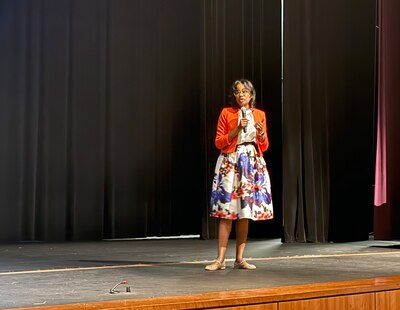 A photograph of a Black woman in a dress on stage speaking into a microphone.