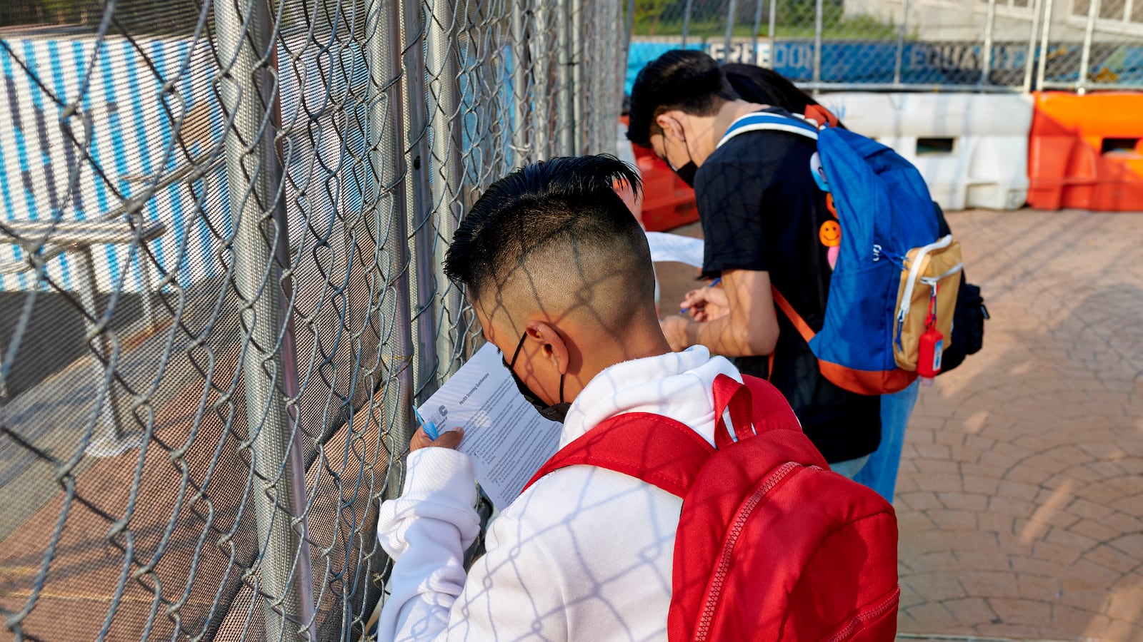 High school students complete their COVID screening forms next to a chainlink fence outside of their school’s entrance.