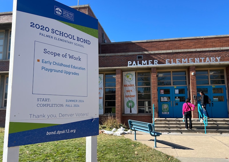 Two people walk into the front entrance to a brick elementary school with a sign above the entrance that reads "Palmer Elementary." There is a white sign in the grass that has blue and black text on it.