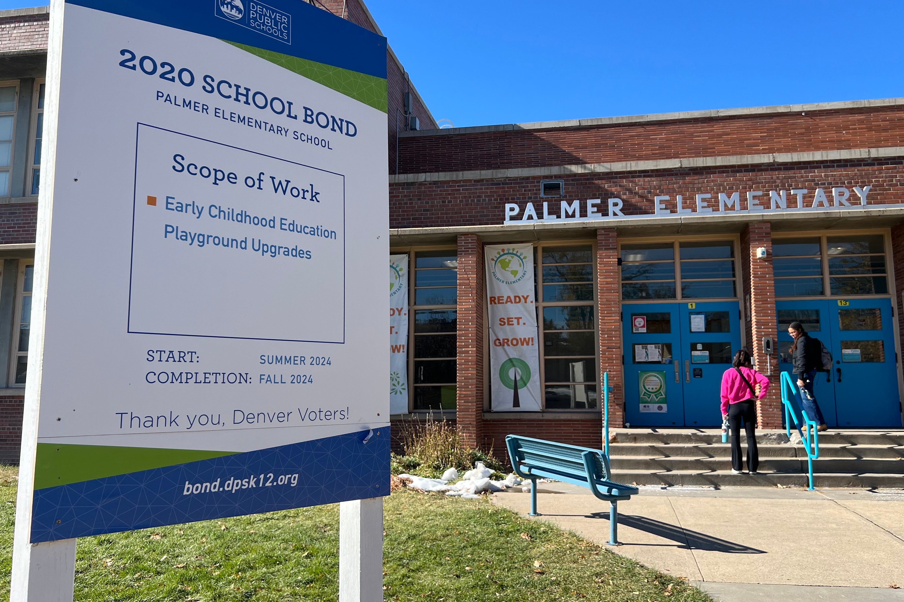 Two people walk into the front entrance to a brick elementary school with a sign above the entrance that reads "Palmer Elementary." There is a white sign in the grass that has blue and black text on it.