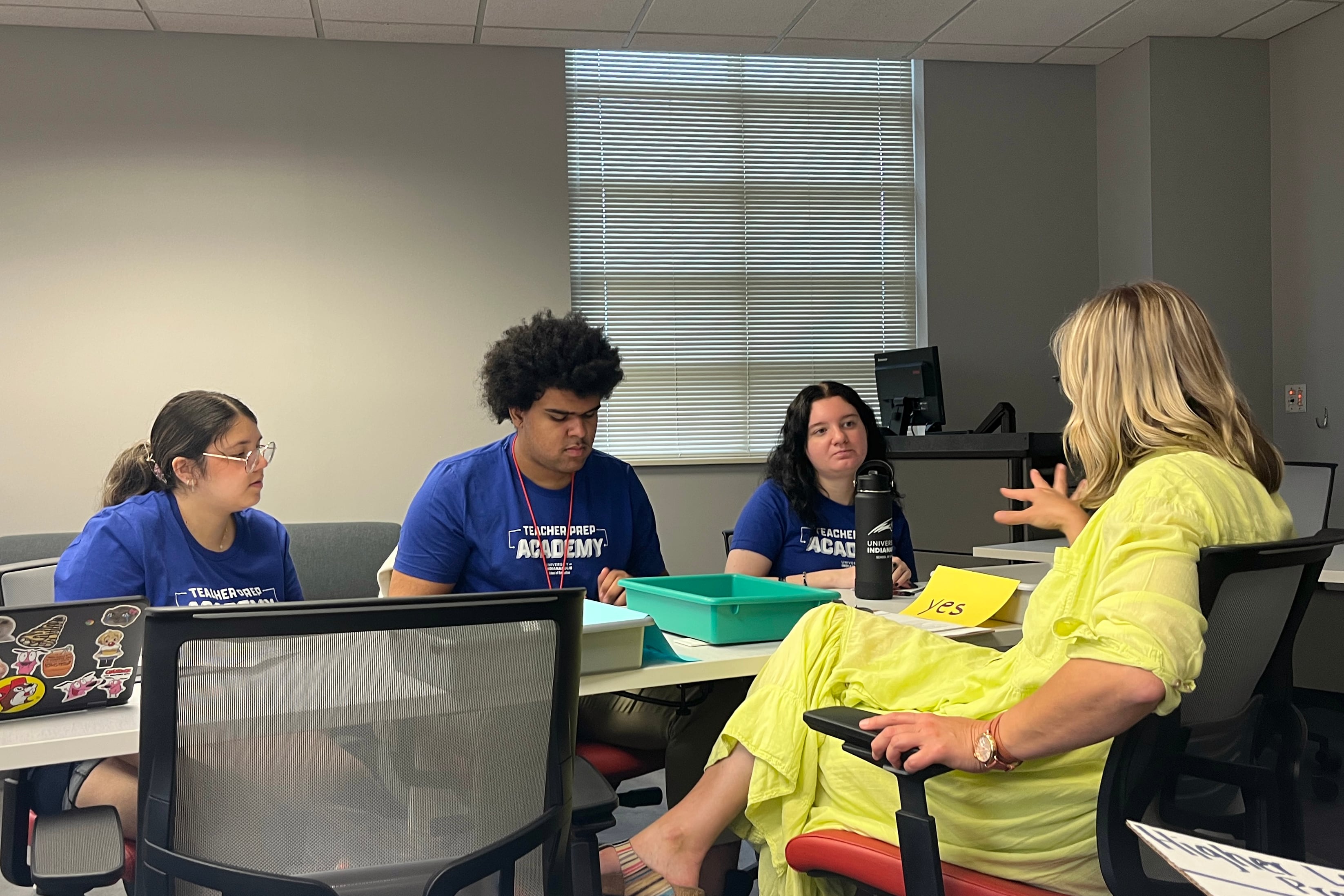 Three young adults in blue t-shirts sit on one side of a table while one adult in a yellow dress sits on the other side.
