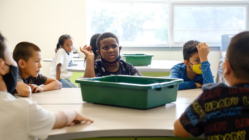 Elementary students with and without masks sit around a table with a green plastic bin in the middle of the table.