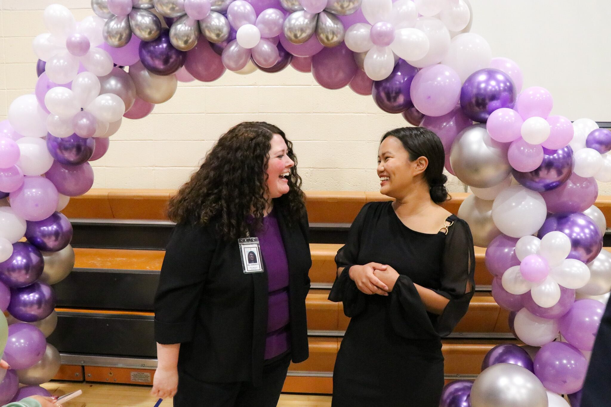 Two women standing under a purple, white, and silver balloon arch in a school gym.