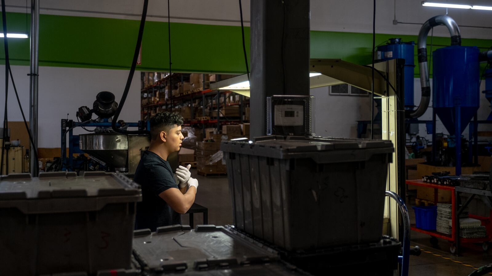 A young man puts a pair of latex gloves on as he prepares for a shift at a factory. The factory lights hanging from the ceiling illuminate him amongst several boxes and pieces of machinery.