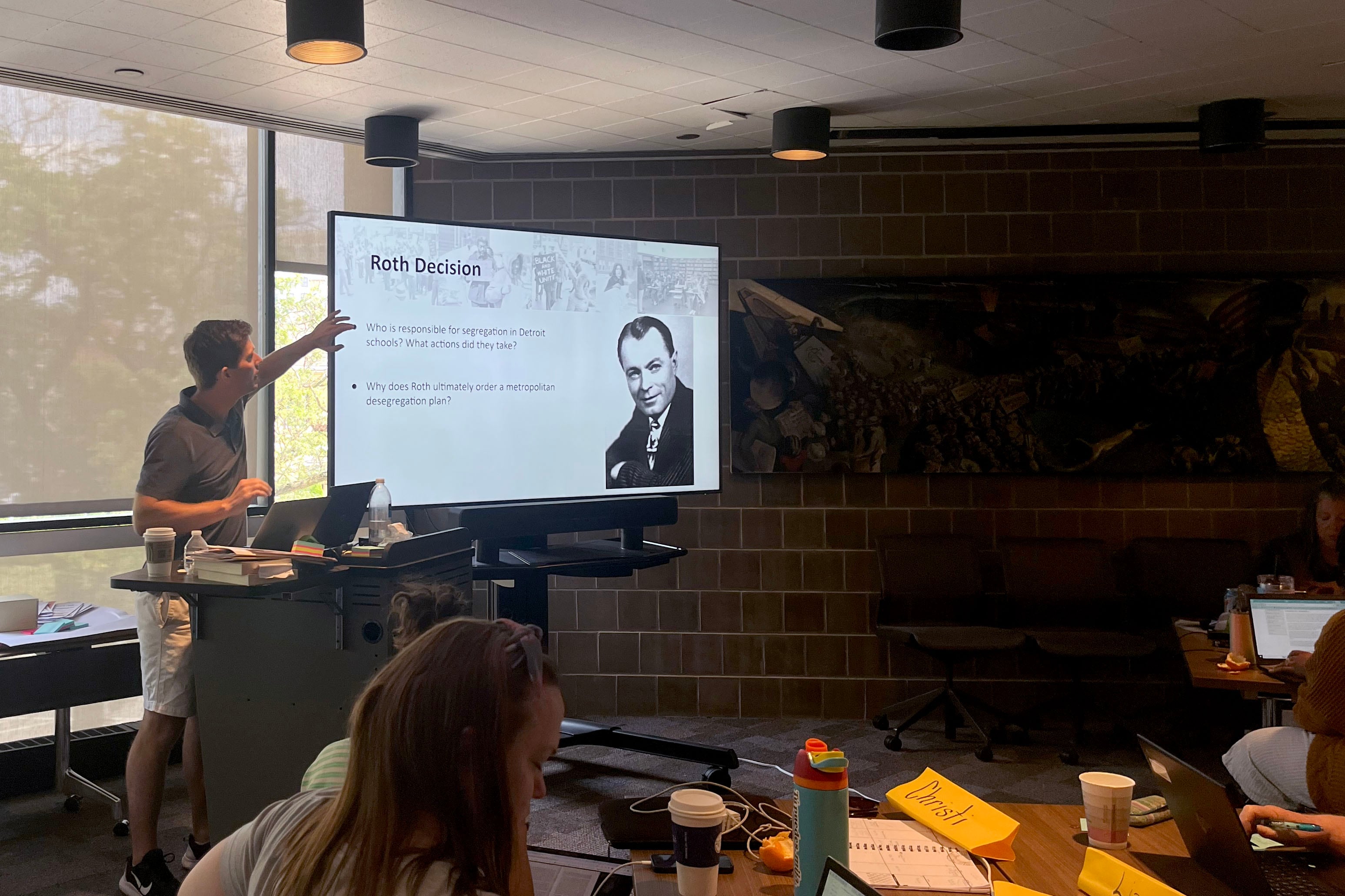 A man speaks from a podium next to a large screen and in front of a group of people sitting at tables spread across a conference room.