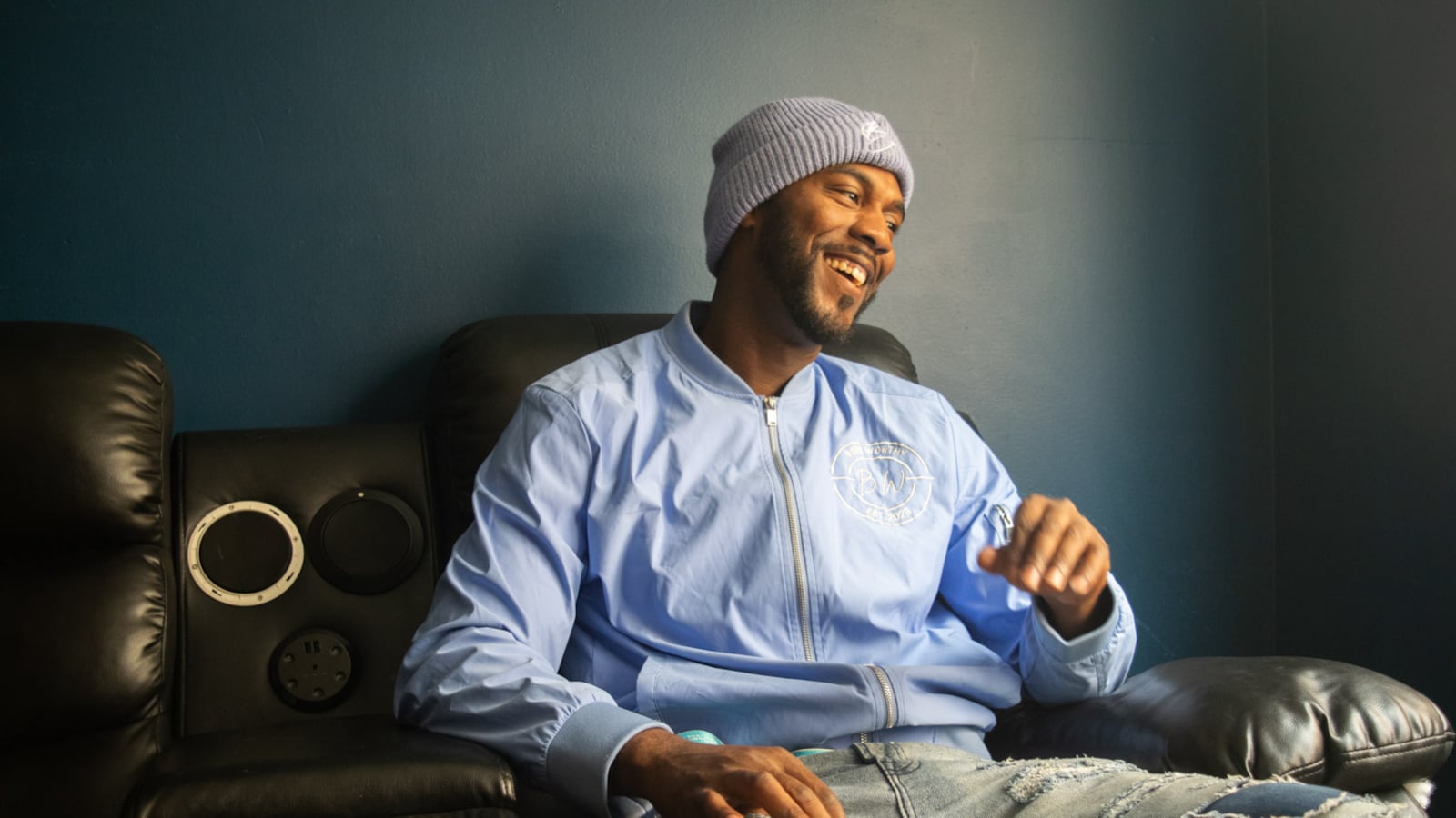 A man sitting on a black couch smiles and looks off into the distance, his eyes slightly down. He is turned toward a window, so light illuminates his body. He is wearing a knit beanie, blue windbreaker, and lightwash jeans. The jeans have rips with a darker denim underneath them. One of his hands is mid-motion, coming down from him rubbing his mouth and laughing. He looks comfortable and laid back.