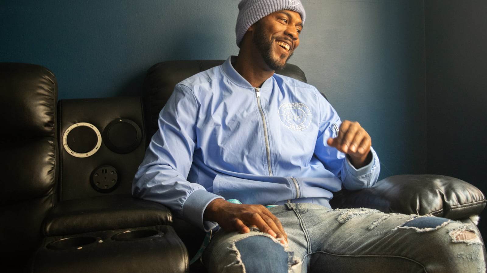 A man sitting on a black couch smiles and looks off into the distance, his eyes slightly down. He is turned toward a window, so light illuminates his body. He is wearing a knit beanie, blue windbreaker, and lightwash jeans. The jeans have rips with a darker denim underneath them. One of his hands is mid-motion, coming down from him rubbing his mouth and laughing. He looks comfortable and laid back.