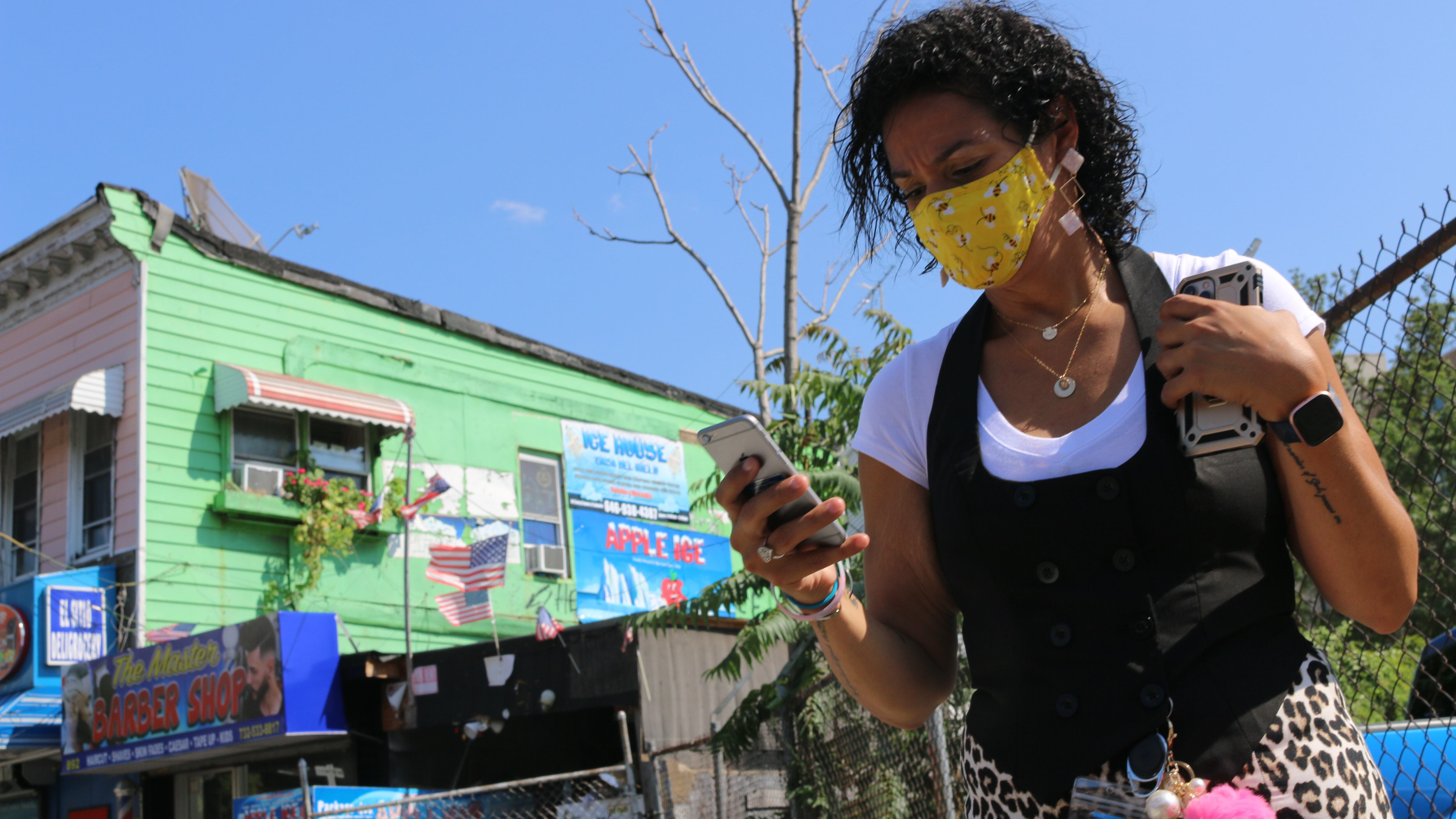 A woman in a yellow mask looks at her phone, with a green building behind her.