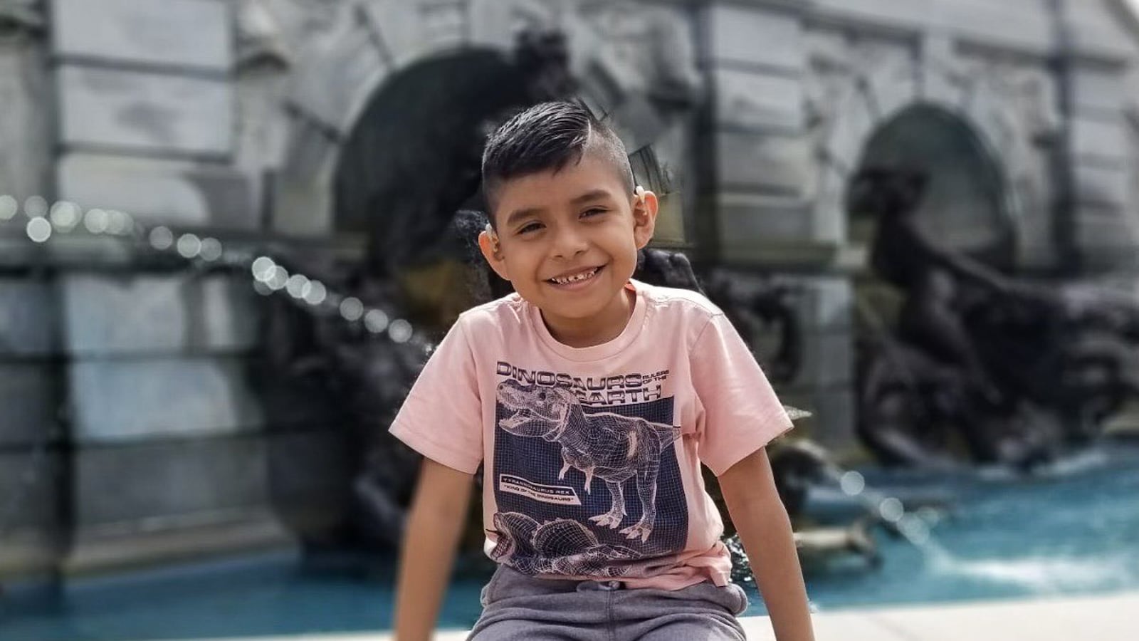 A third grade boy sits at the edge of a fountain, smiling toward the camera.
