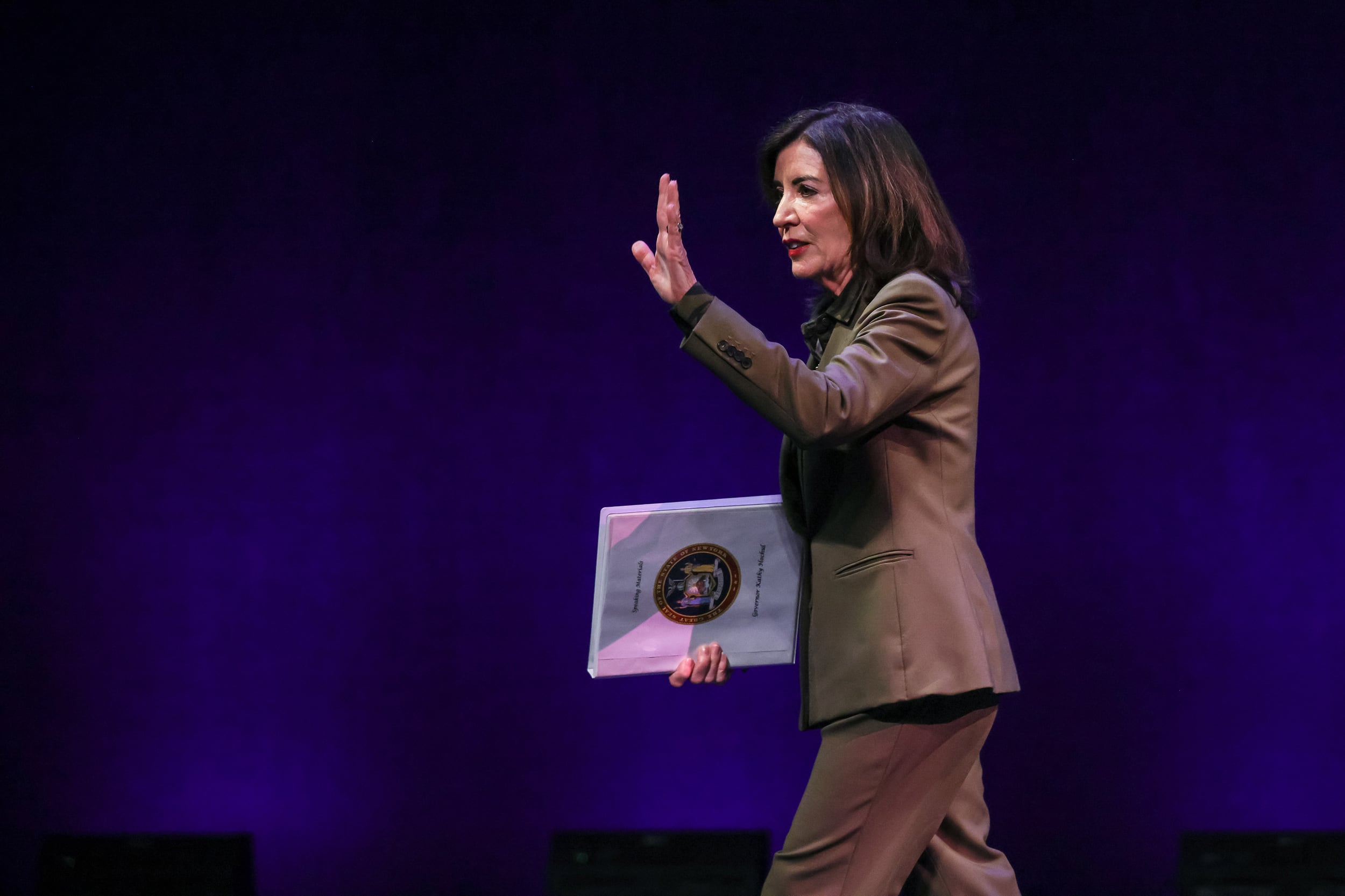 A photograph of a white woman in a suit holding paper while waving and walking across a stage.