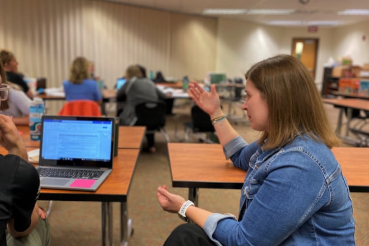 A woman in a jean jacket sits at a desk and talks as a man sits next to her in a black shirt and glasses with a laptop computer in front of him.